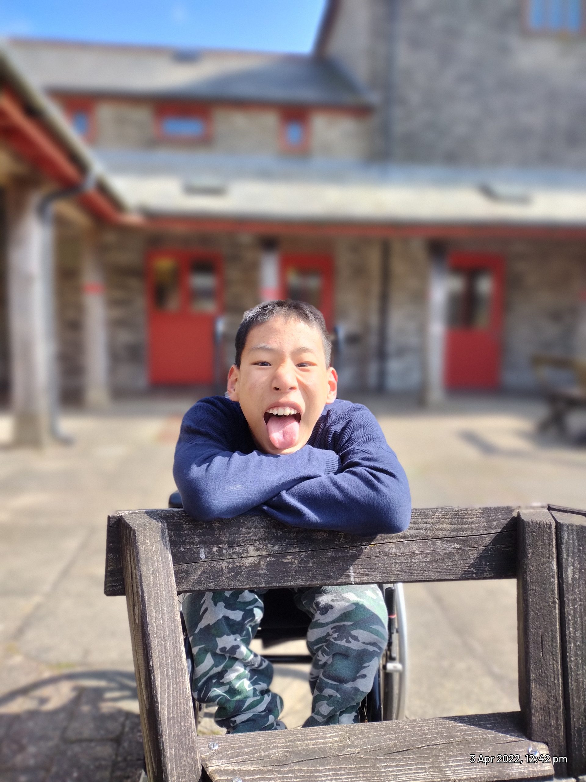 A boy wearing a blue sweatshirt and camouflaged trousers is leaning on the back of a bench and sticking his tongue out at the camera.