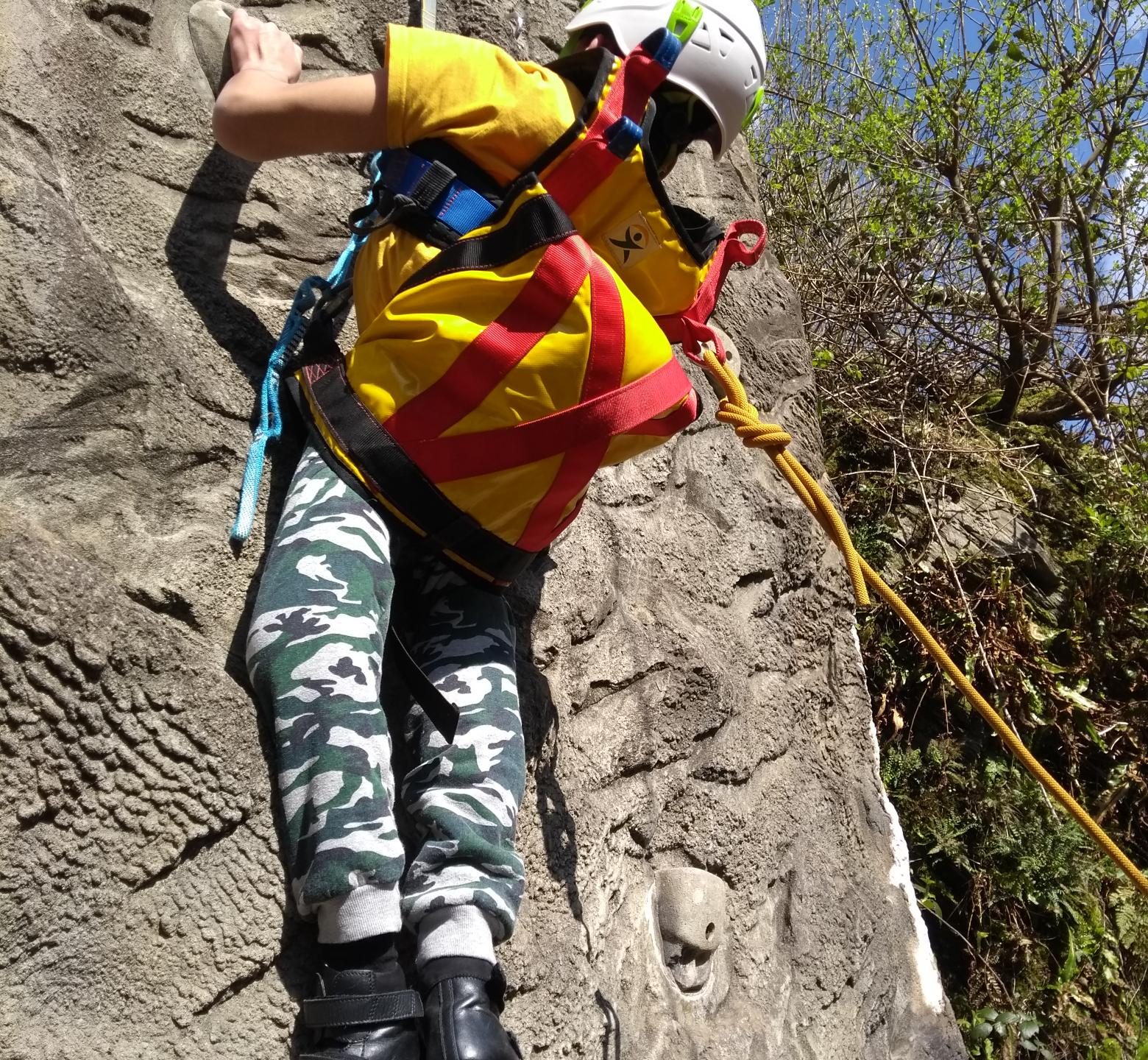 A guest is suspended by ropes on the outdoor climbing wall, wearing a yellow sling. His legs are hanging down below him in camouflage trousers.