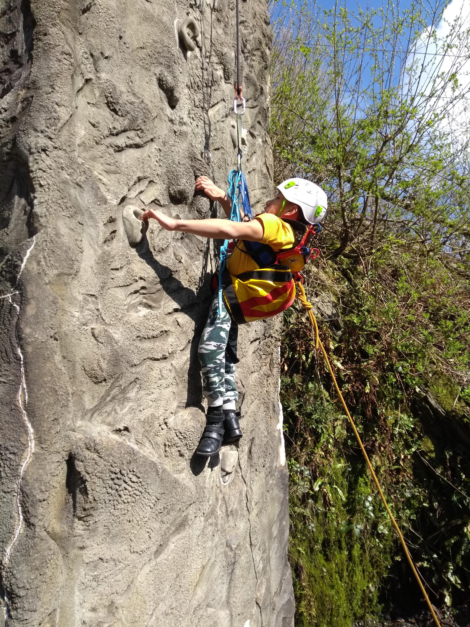 A guest wearing a white helmet and a yellow sling harness is suspended in front of an outdoor climbing wall. Their hands are holding some climbing holds and their legs are hanging below them.