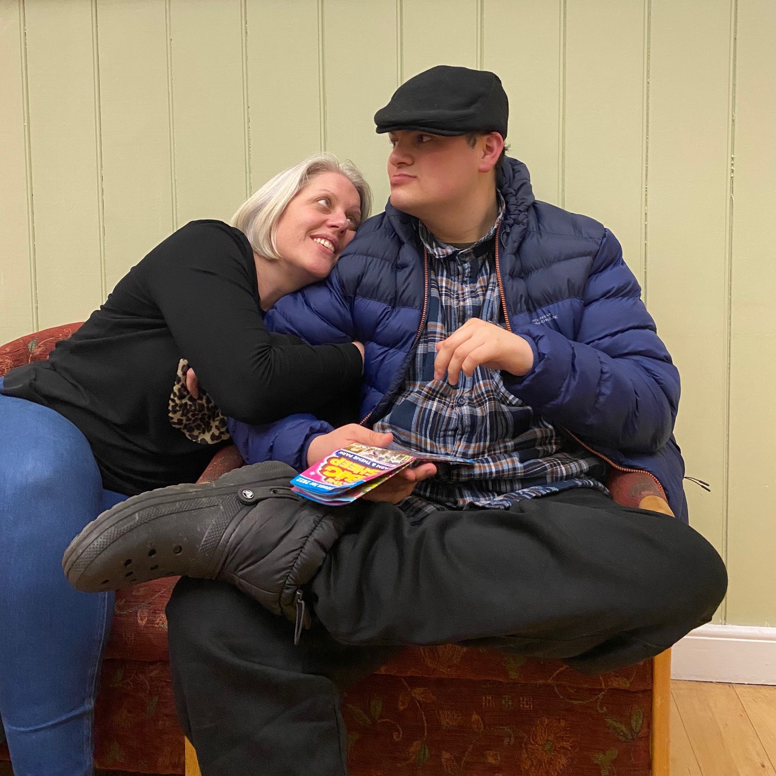 A man is sitting looking away from the camera, wearing a hat and coat, with his leg crossed across the other. He is holding a leaflet and on his arm, is a woman's arm, as she leans her head on his shoulder looking up at him in the chair next to him.