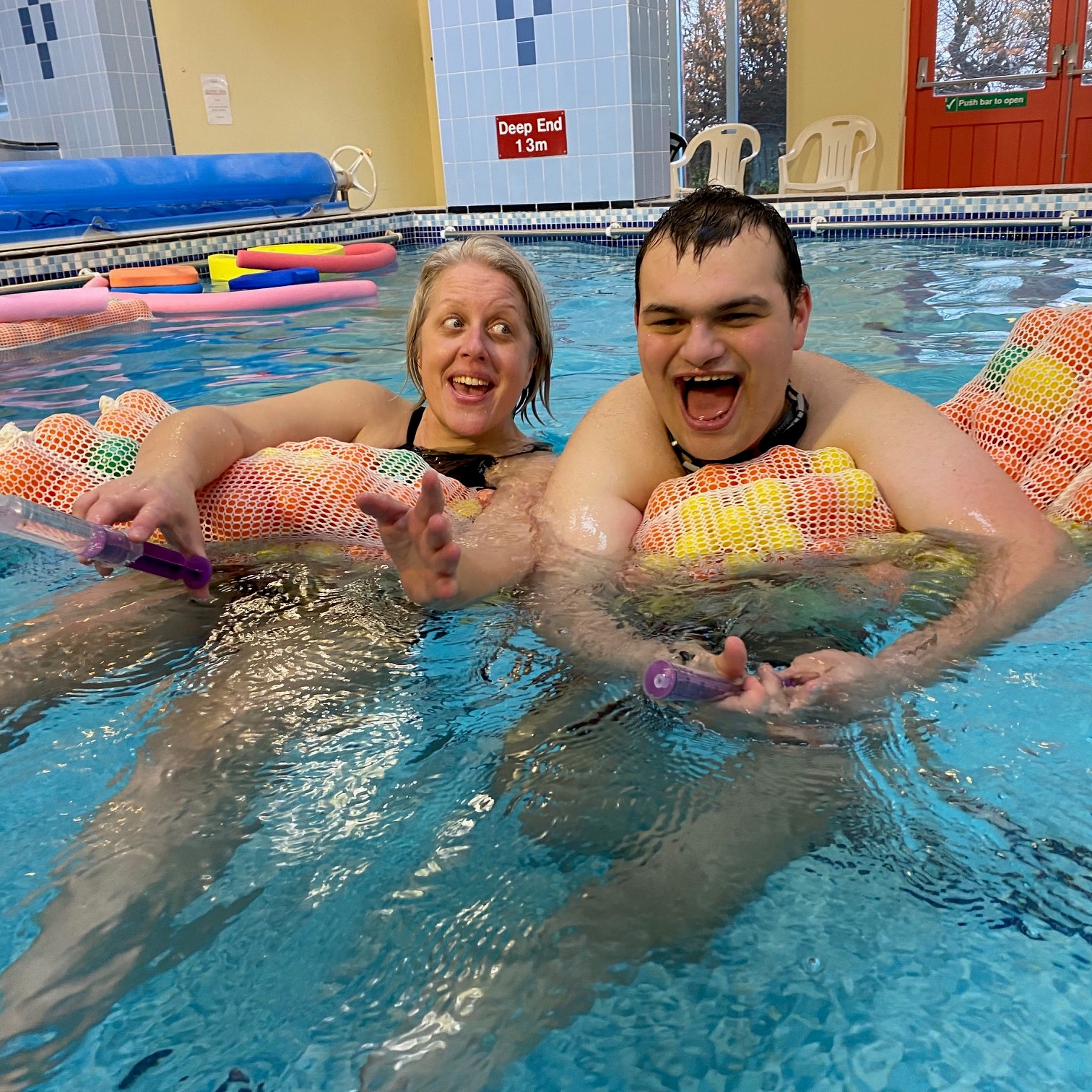 A woman and a man are holding onto a floating netted bag in a blue swimming pool and smiling mouths open at the camera.