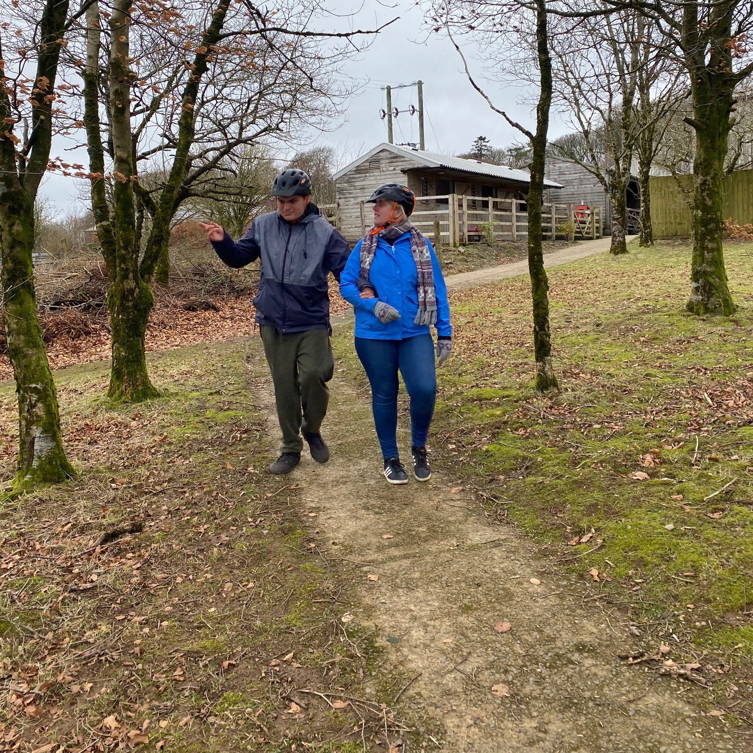 Two guests at Calvert Exmoor are walking down a path through woodlands wearing bike helmets. One guest has his arm resting on the arm of the woman next to him as they are walking.