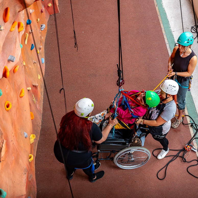 An aerial view of a guest in a wheelchair being hoisted up by ropes in a sling with the help of 3 guests in helmets around the wheelchair user next to the indoor climbing wall.