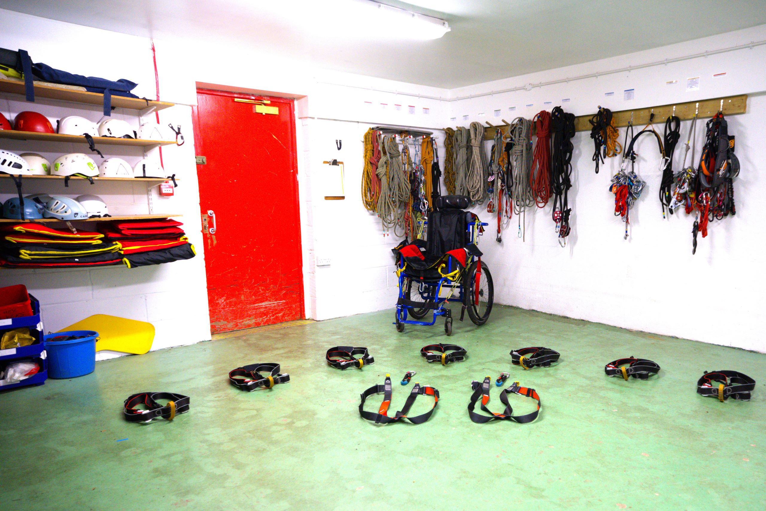 A room with a red door and green floor. There are helmets on shelves and ropes hanging on hooks on the wall, a wheelchair in the centre at the back and in the foreground, there is a selection of harnesses on the floor laid out ready for guests to put them on.