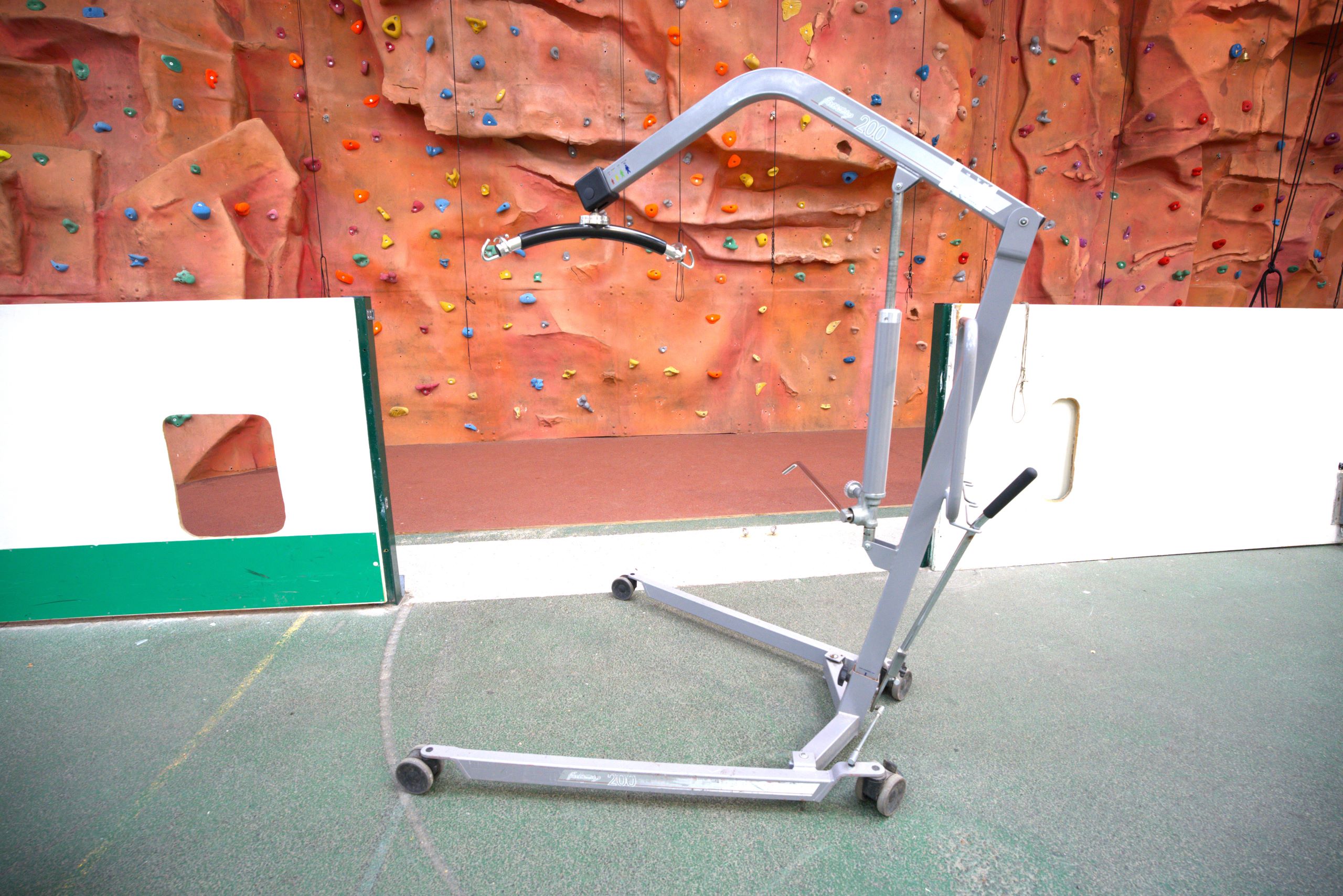 A silver metal mobile hoist in front of the climbing wall with a black T section with hooks on.