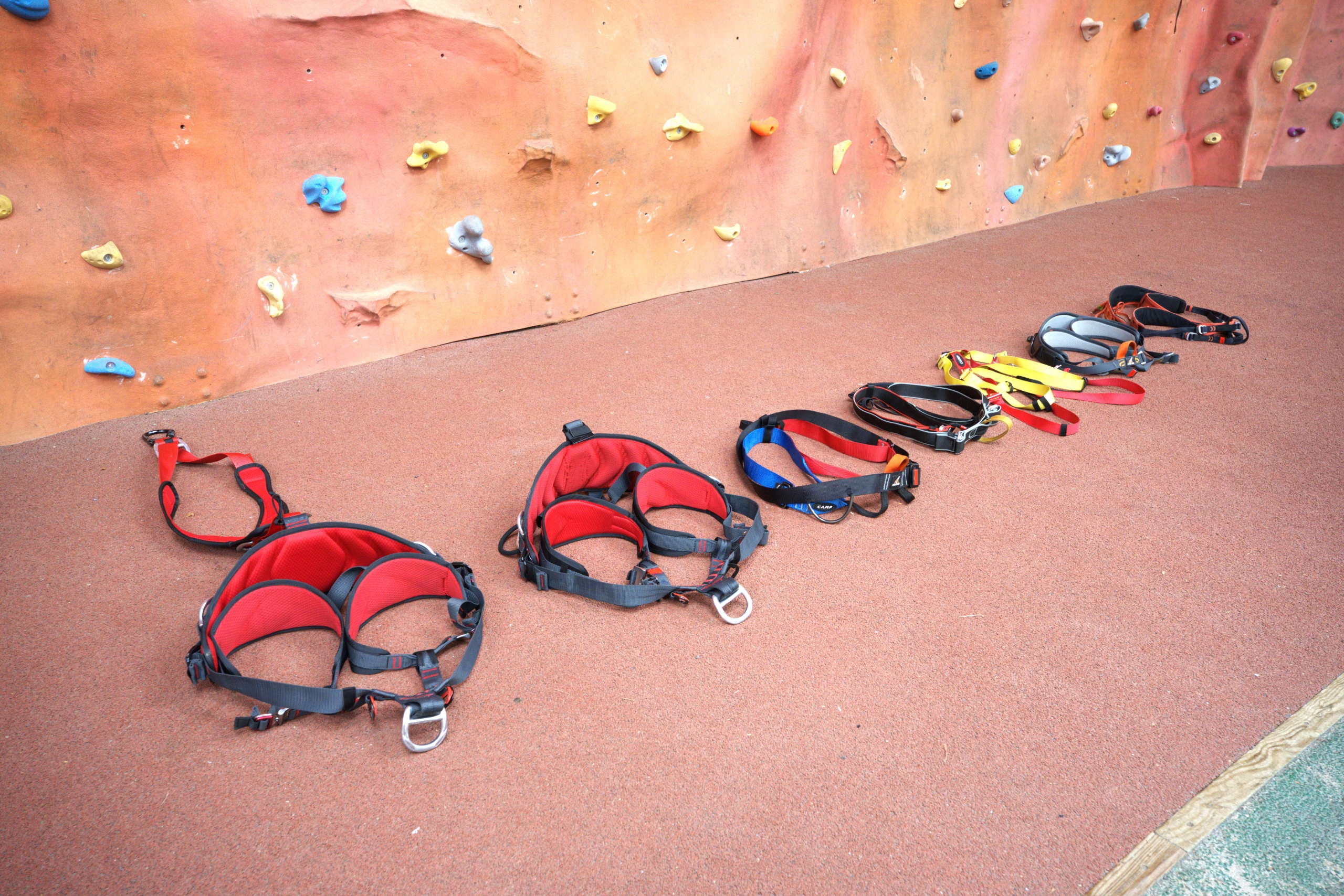 A selection of seven different harnesses laying out on the floor at the bottom of the indoor climbing wall.