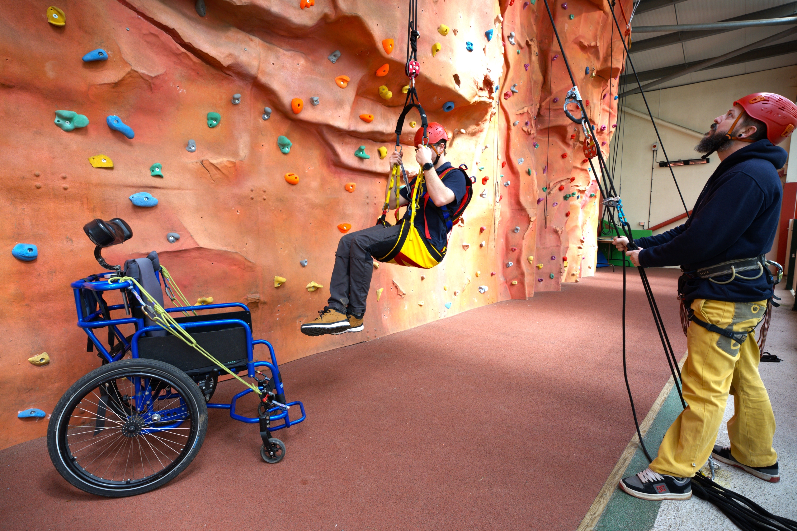 A member of Calvert Exmoor staff is sitting in the accessible sling and being suspended above the ground by another member of staff who is using a belay system in the foreground. The blue abseiling chair is to the left of the picture.
