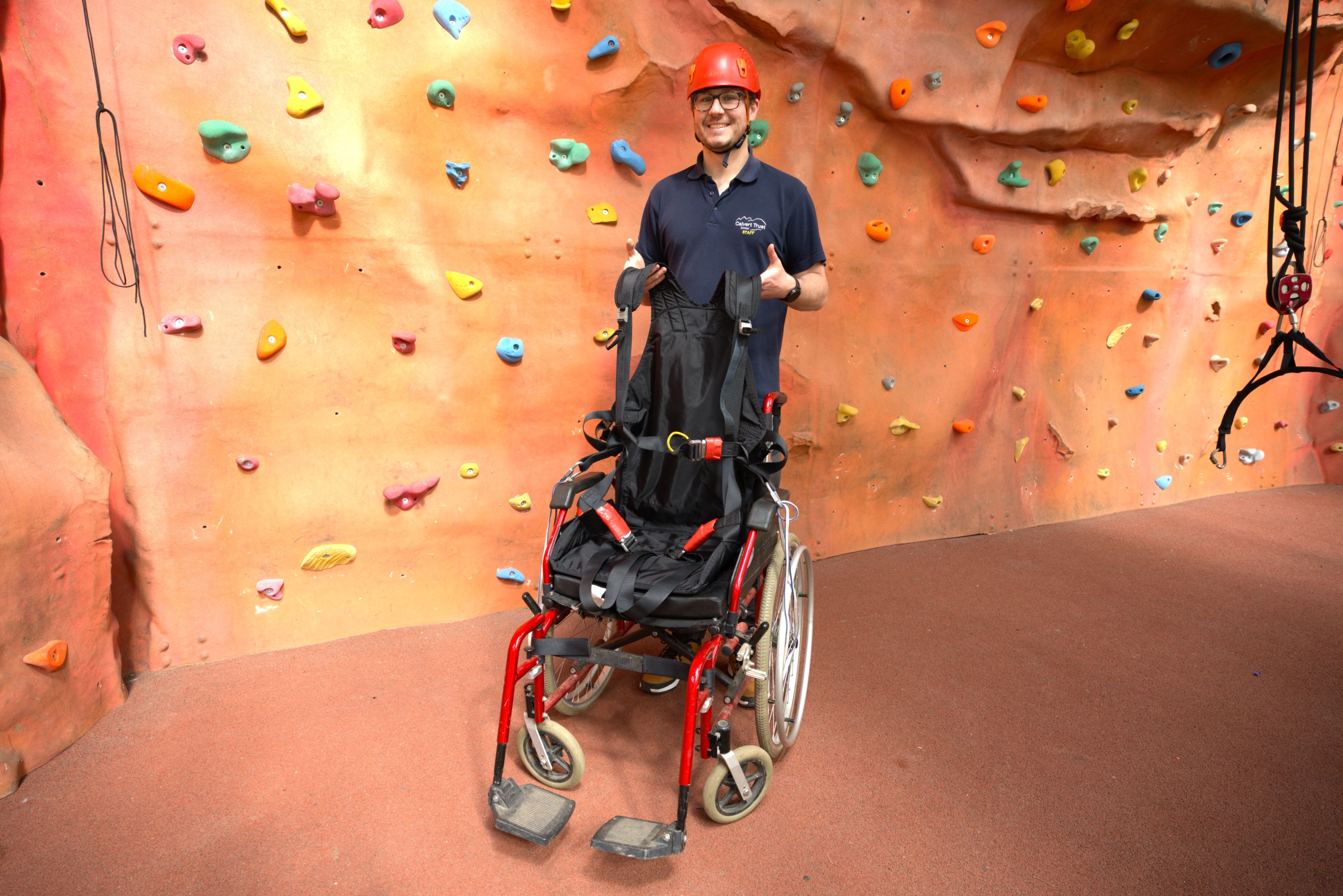 A member of Calvert Exmoor staff smiles at the camera holding up a harness, which can be used for accessible climbing and abseiling, within a red wheelchair in front of the indoor climbing wall.
