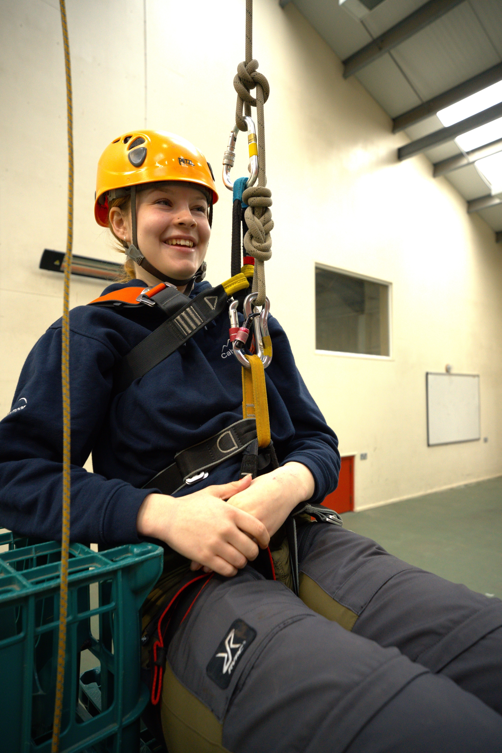 A Calvert Exmoor instructor sitting on some crates smiling, using a sit and chest harness, attached into a rope system with her hands in her lap.