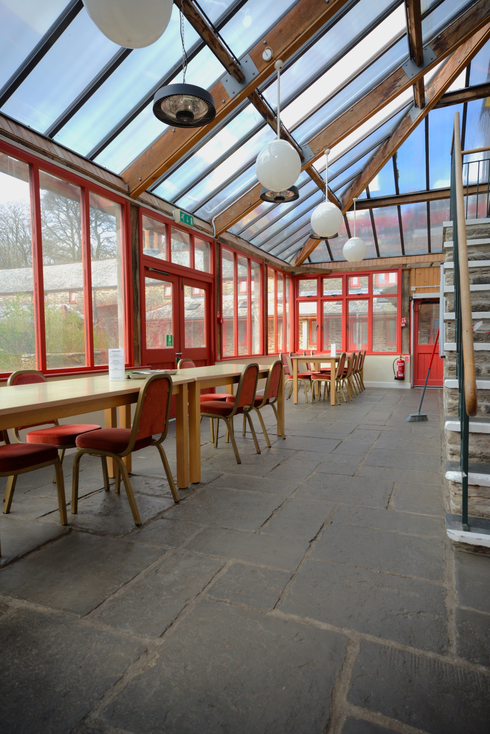 A naturally lit conservatory room, with a set of stairs to the right and tables with chairs on the left set up like a dining room.