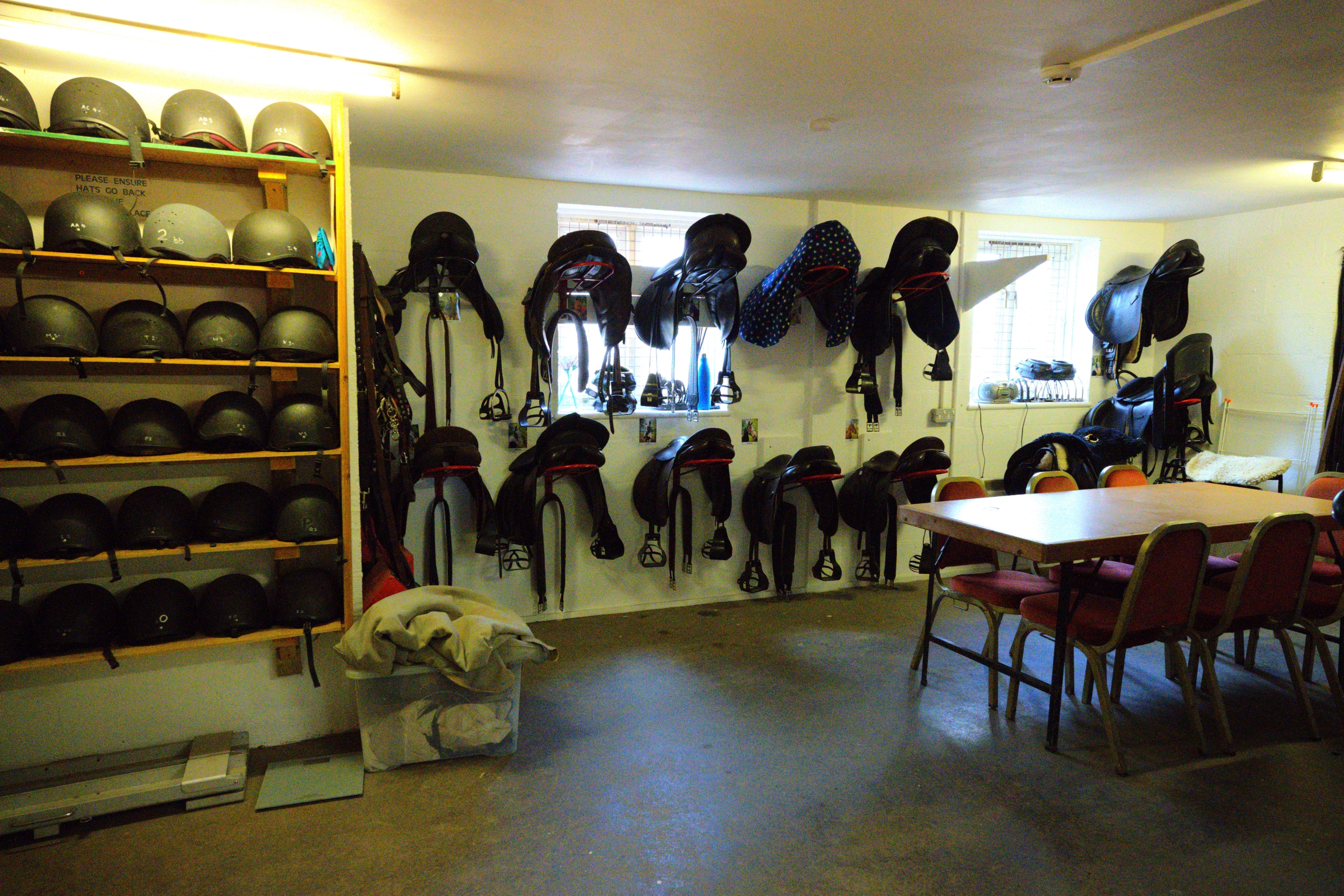 The tack room in the Equestrian Centre. There's a row of saddles on the back wall, a table with chairs around it in front and then to the left, shelves filled with riding hats.