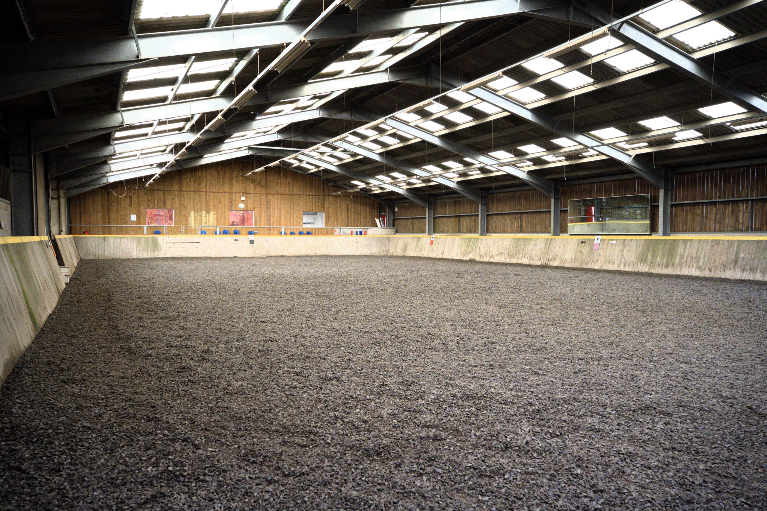 An empty indoor riding arena, with a floor of black rubber shavings, wooden walls and metal roofing with natural light. At the far end is a row of blue chairs in the viewing gallery.