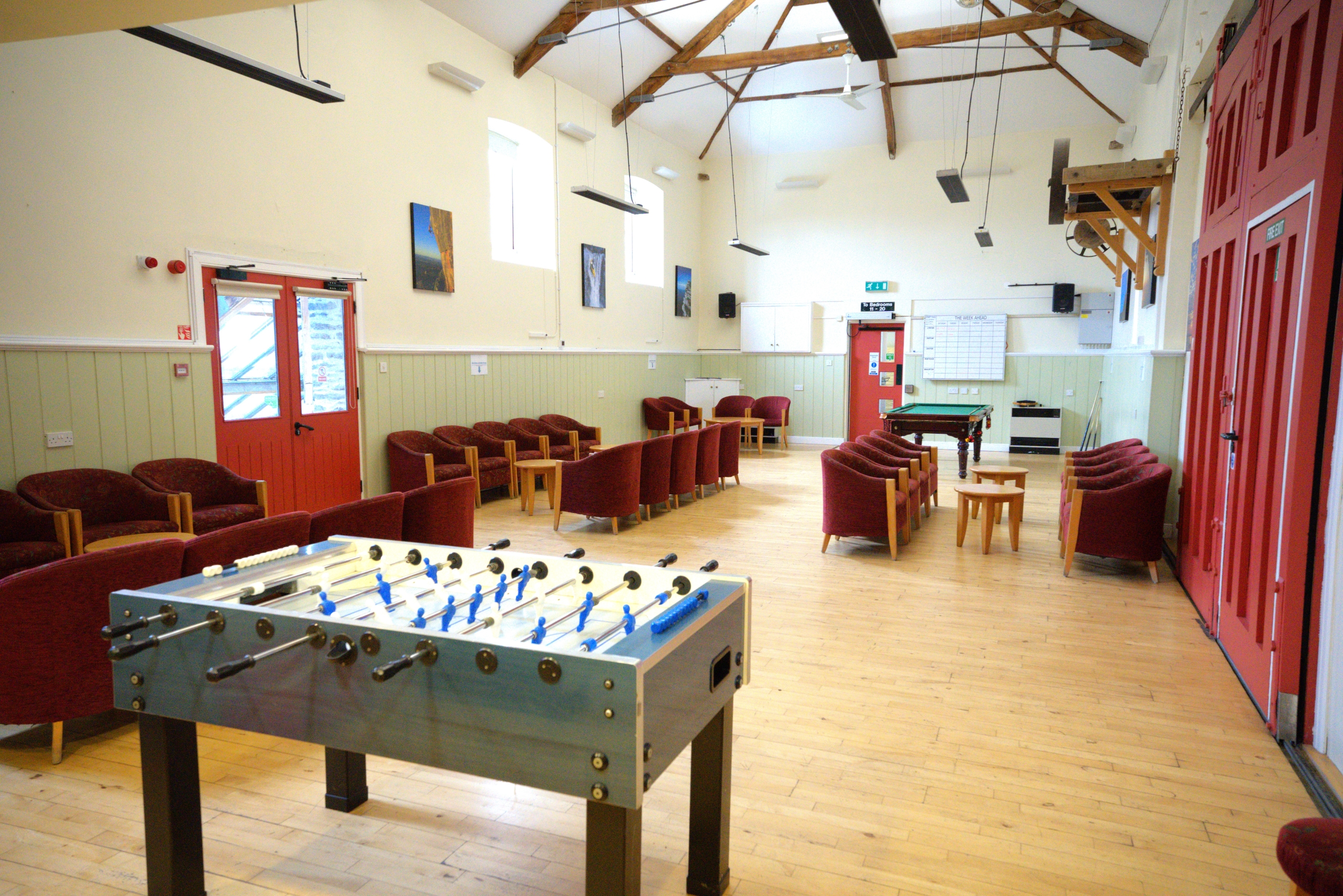 An open plan naturally lit indoor barn conversion with red doors to the right, one on the left and one at the back of the room with rows of comfortable arm chairs and tables, as well as a pool table and indoor foosball table.