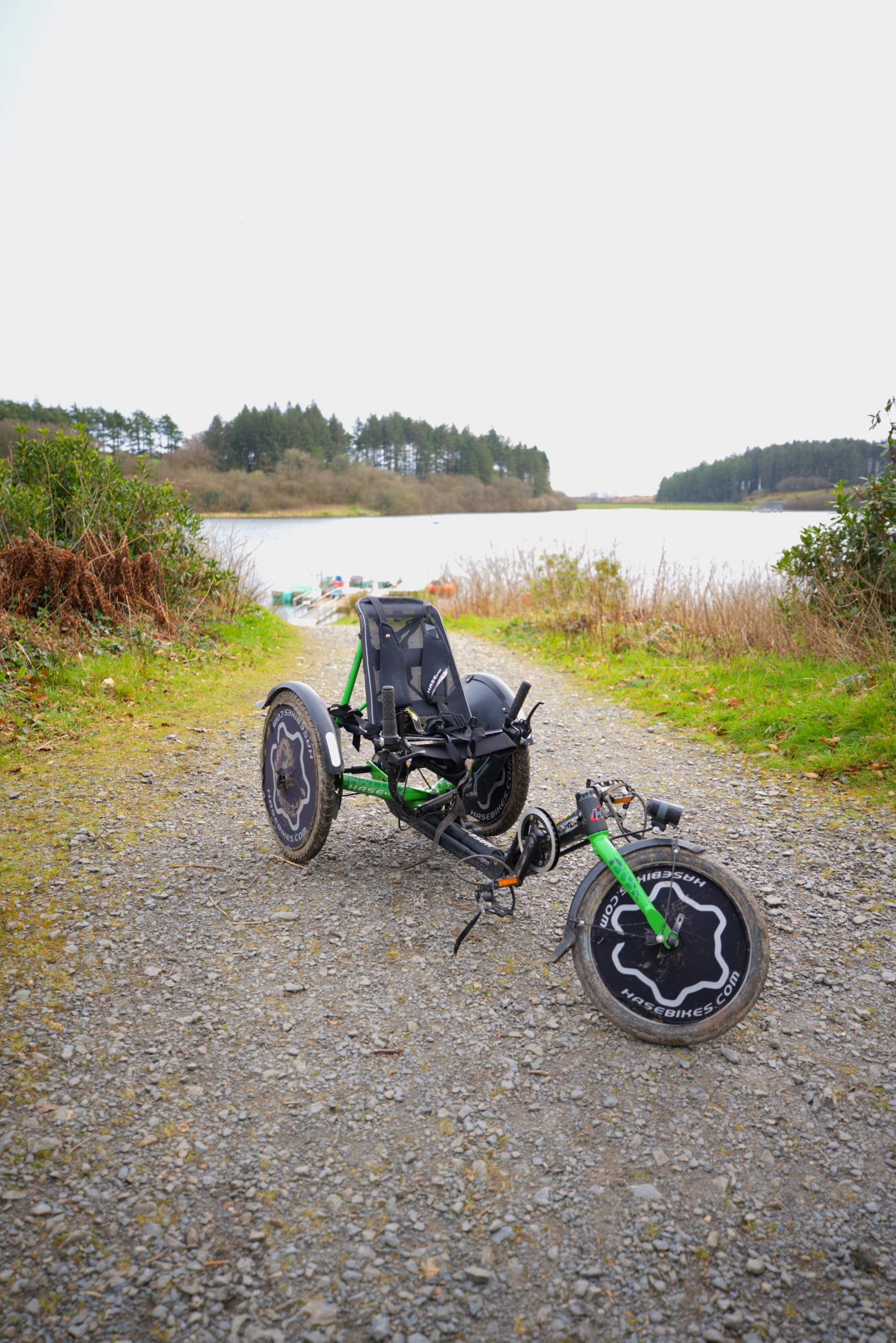 A white accessible trike bike facing the camera on a gravel path that leads down to the reservoir.