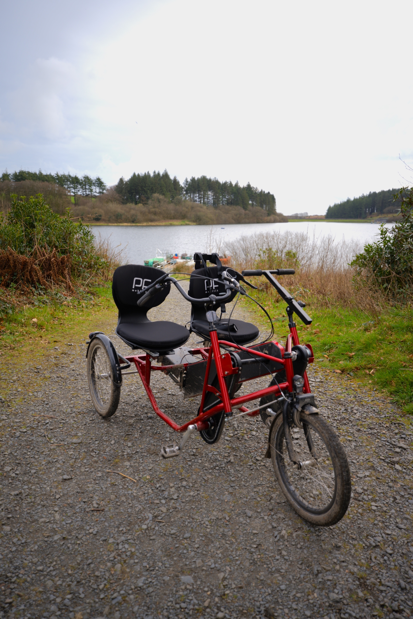 A white accessible trike bike with two seats side by side facing the camera on a gravel path that leads down to the reservoir.