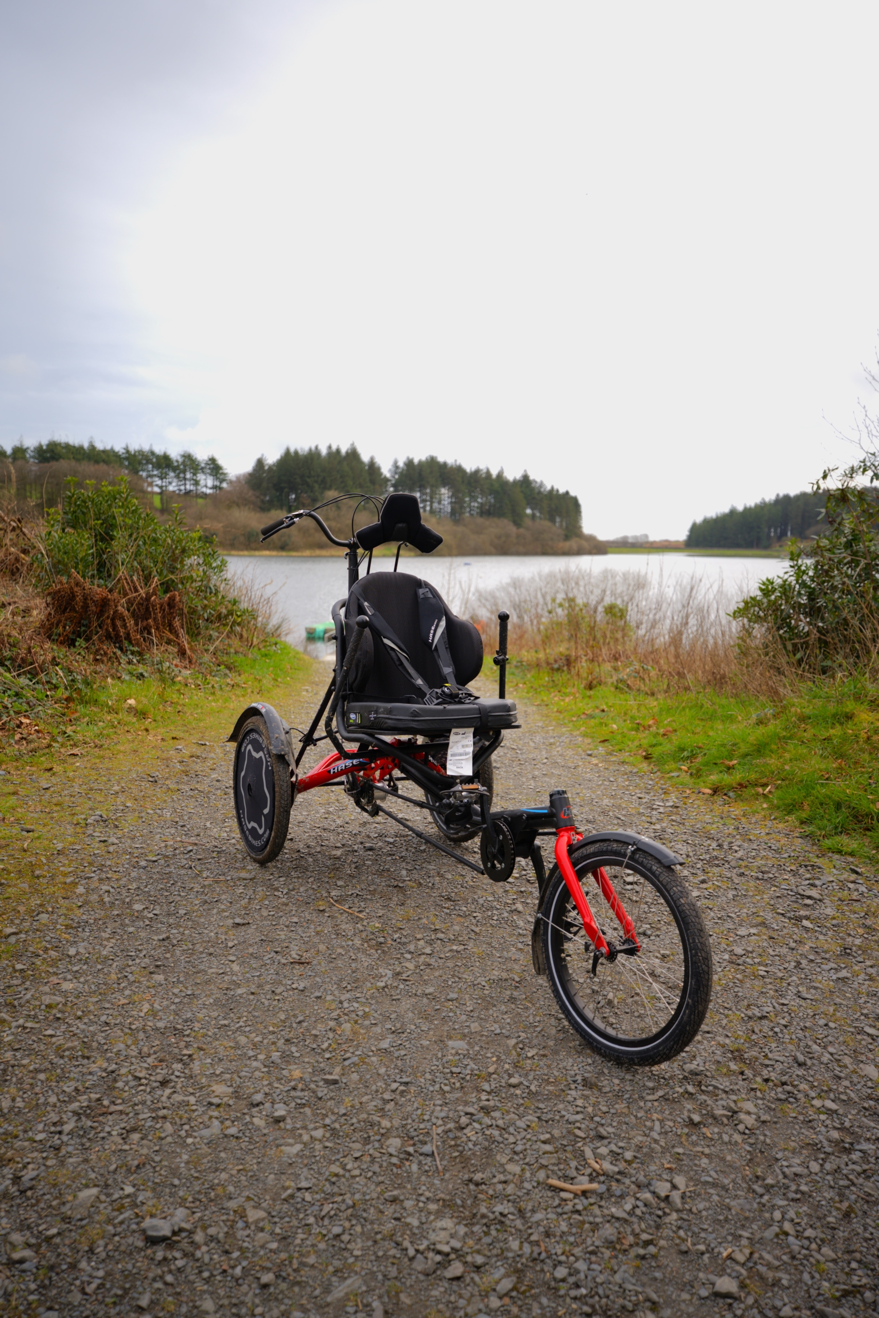 A white accessible trike bike facing the camera on a gravel path that leads down to the reservoir.