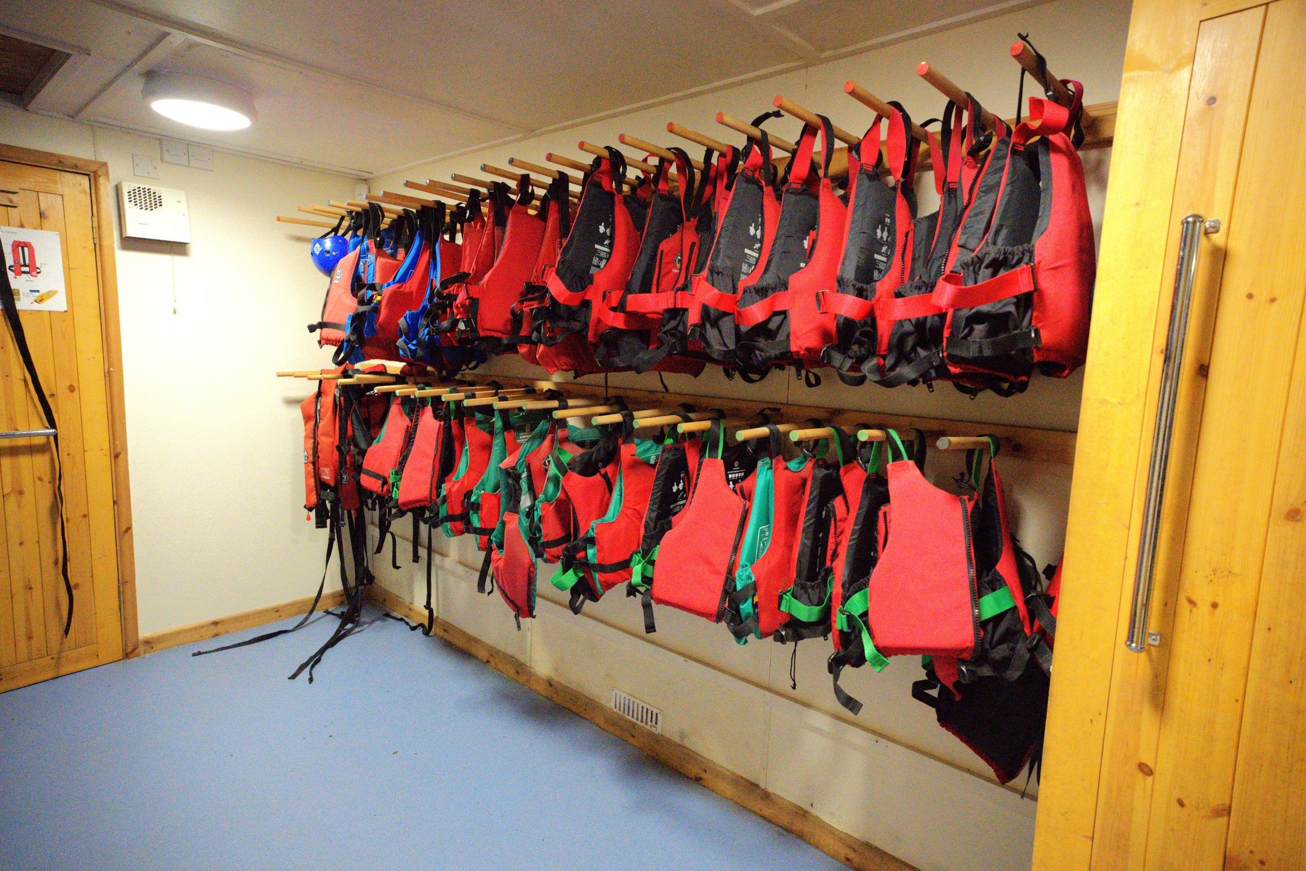 A large selection of red buoyancy aids lined up on pegged shelving in between two wooden doors inside a brightly lit room.
