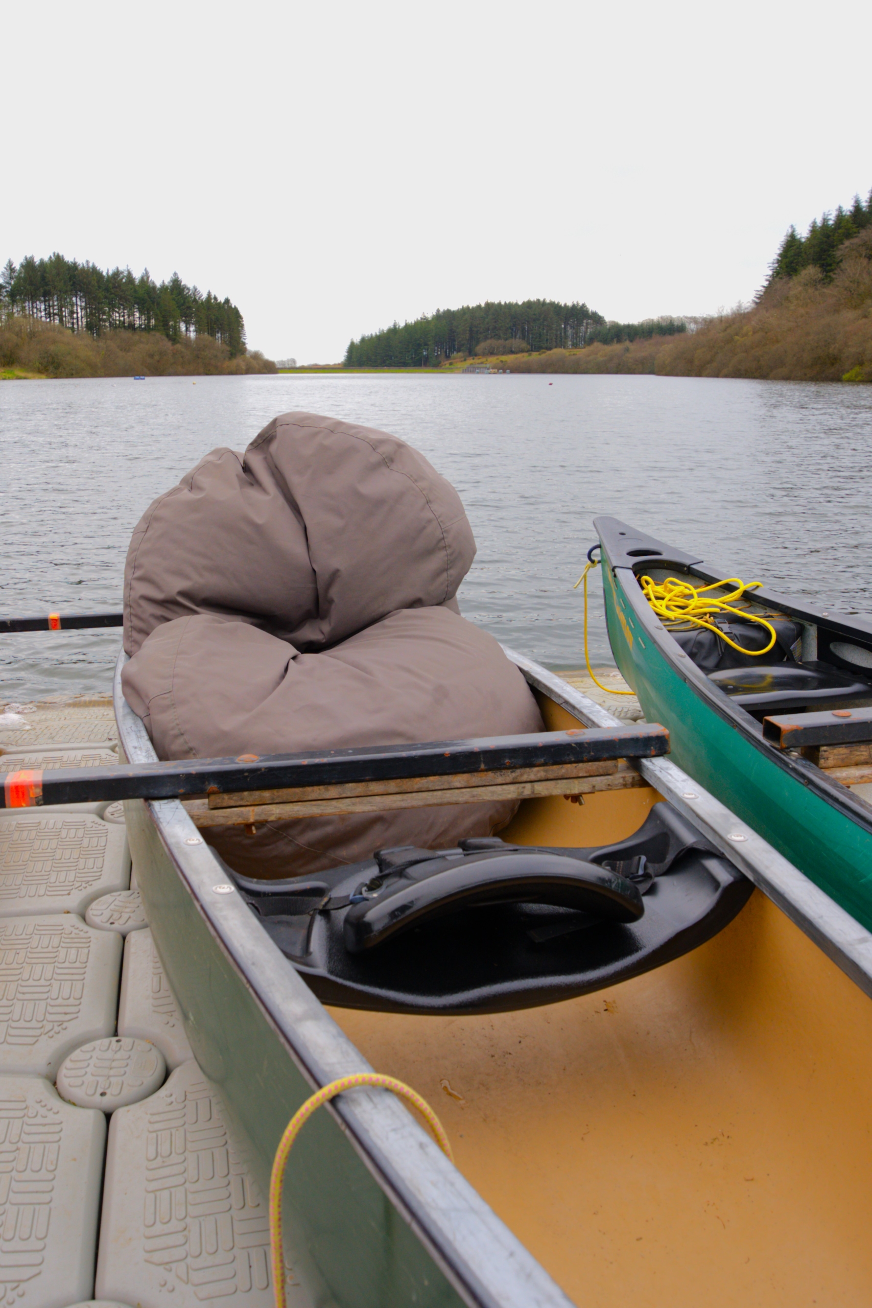 A grey beanbag inside a green and yellow canoe in front of the flat water on a floating jetty.