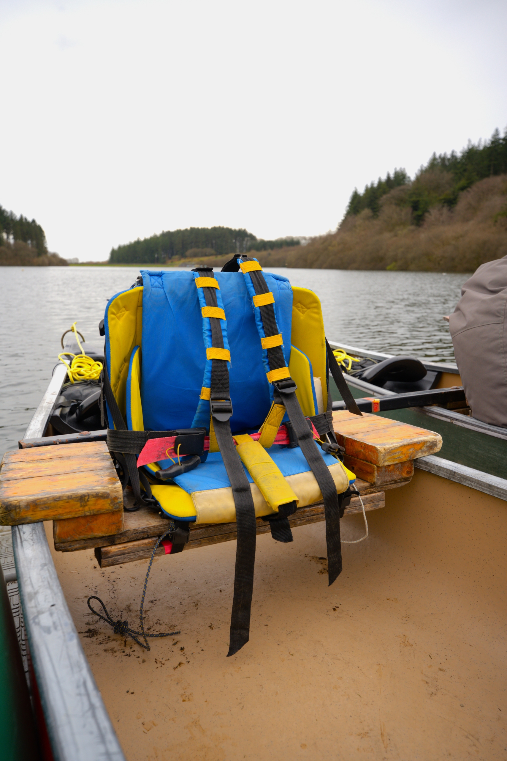 A blue and yellow padded seat with back support and shoulder straps on a wooden seat in a canoe with the reservoir in the background.