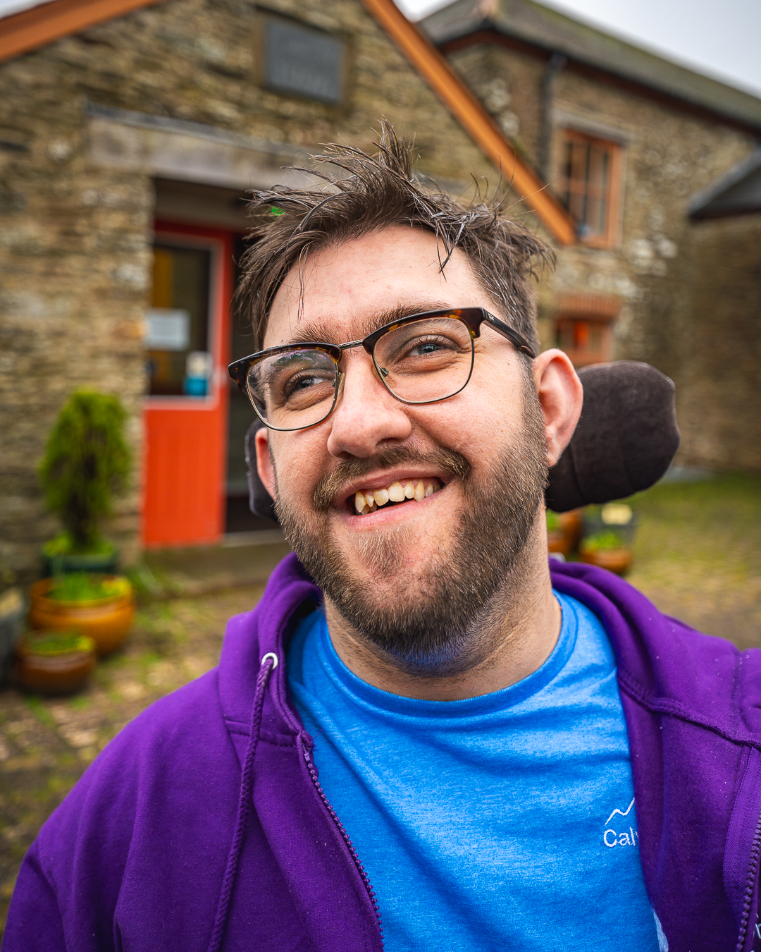 A man with spiky brown hair and a beard, wearing glasses, a blue t-shirt and a purple hooded sweatshirt is smiling at the camera in front of an old stone building.