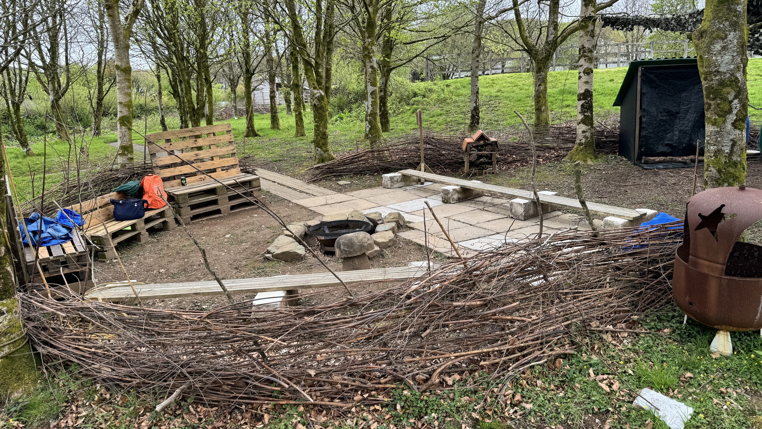 An area within woodlands with woven branches creating a perimeter fencing around. The area has a firebowl in the ground, some benches in a circle shape around the firebowl and off to the right, a shed.