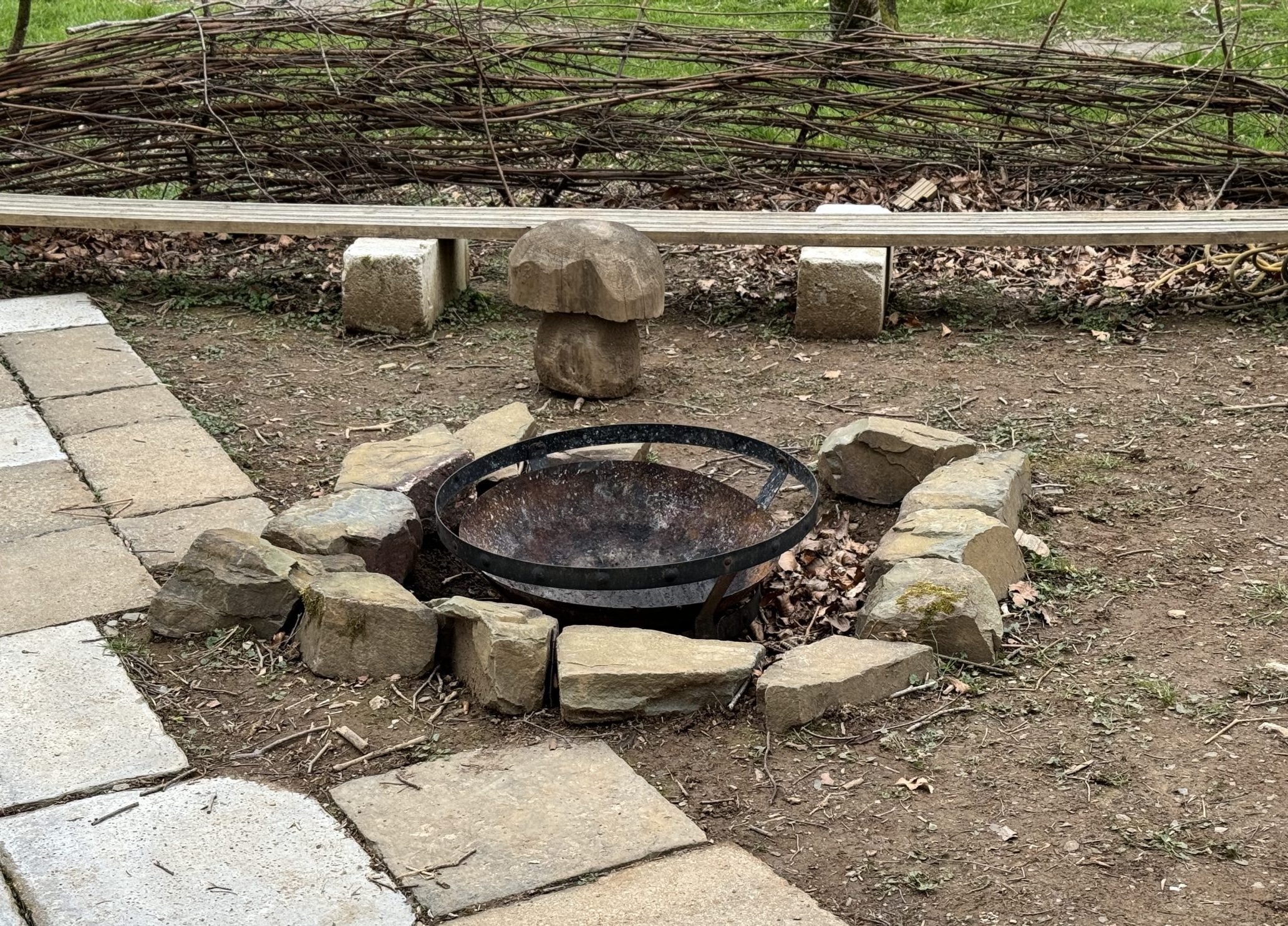 An empty metal fire bowl is in the ground in the accessible bushcraft area. There is paving slabs on the floor on the left of the fire and a bench behind with a woven natural fencing behind it all.
