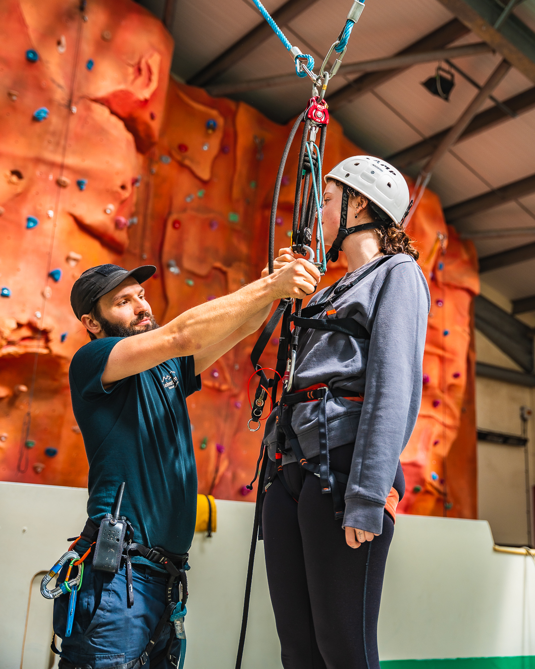 A guest standing in front of the indoor climbing wall with a white helmet and harness, while a member of Calvert Exmoor staff is attaching ropes and carabiners to her harness ready for the Giant Swing activity.