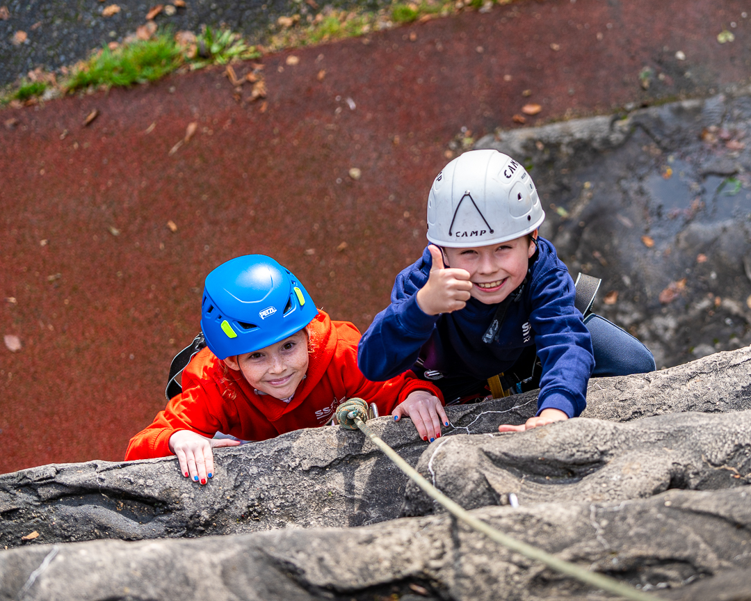 Two children smile looking upwards at the camera above them as they climb the outdoor climbing wall, wearing a blue and grey helmet and harnesses.