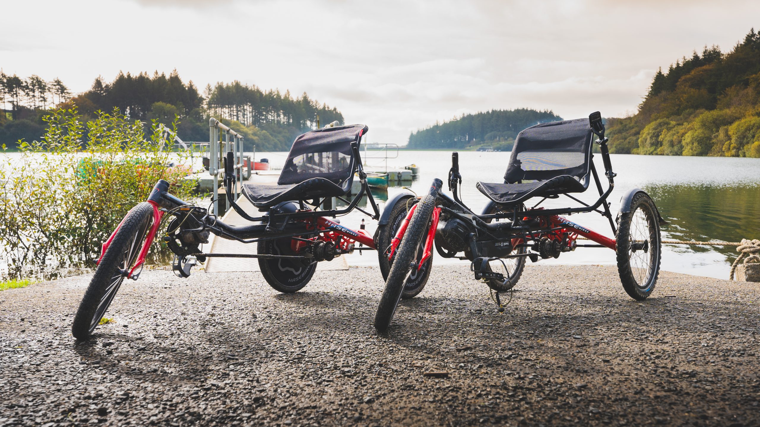 Two red trike bikes are stationary side by side in front of calm water. They have black seats and handlebars either side of the seats.