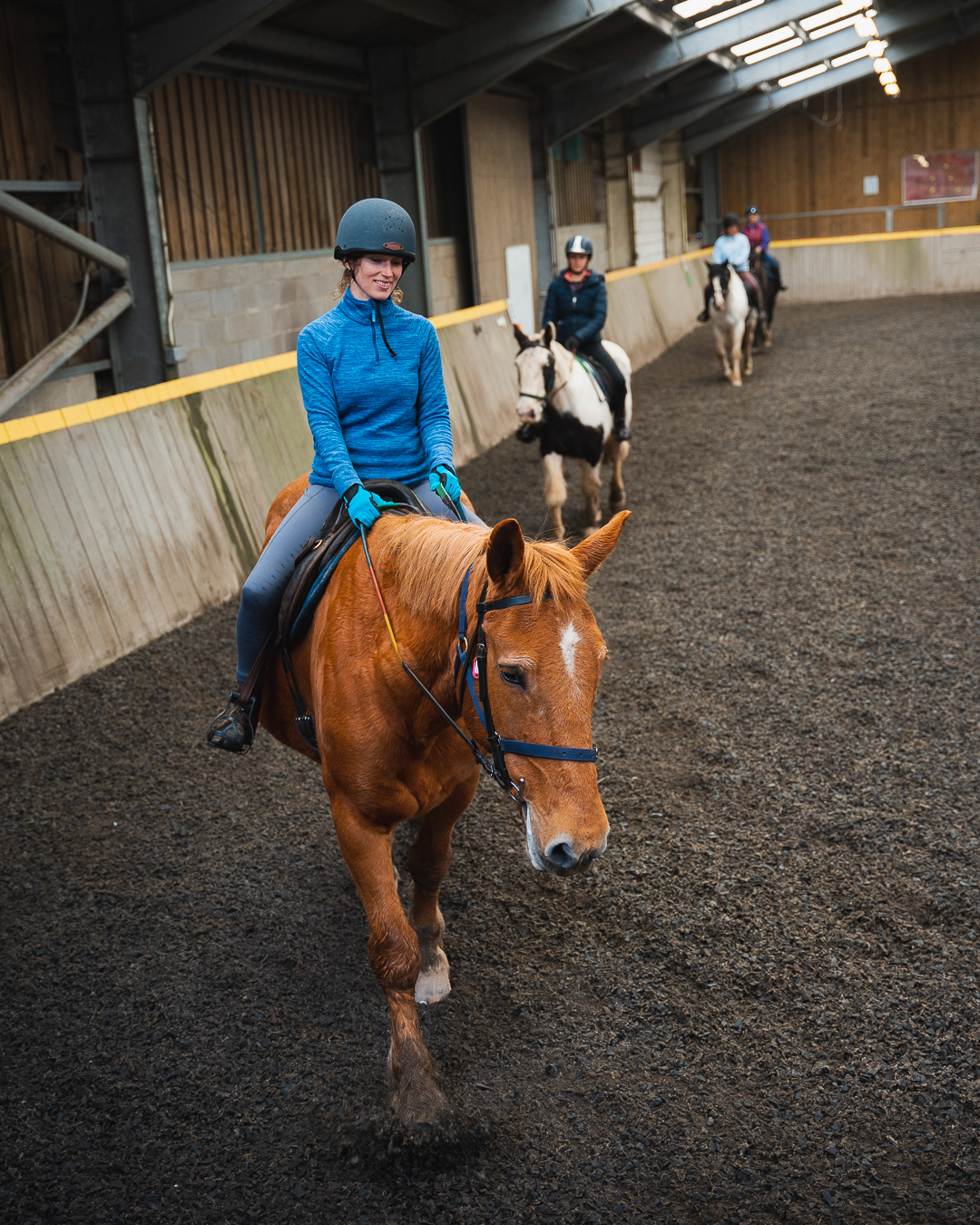A guest in a blue fleece and gloves wearing a riding hat is riding a ginger coloured horse in the indoor arena at Calvert Exmoor's Equestrian Centre. There are other guests on horses in the background.