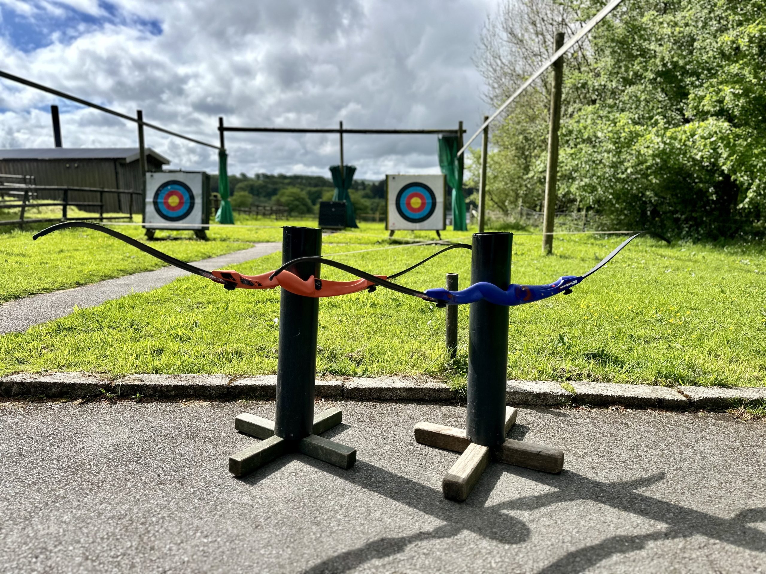 A red and a blue handled archery bow are hanging off two black posts standing on the ground. In the background are two archery targets and grass in between.