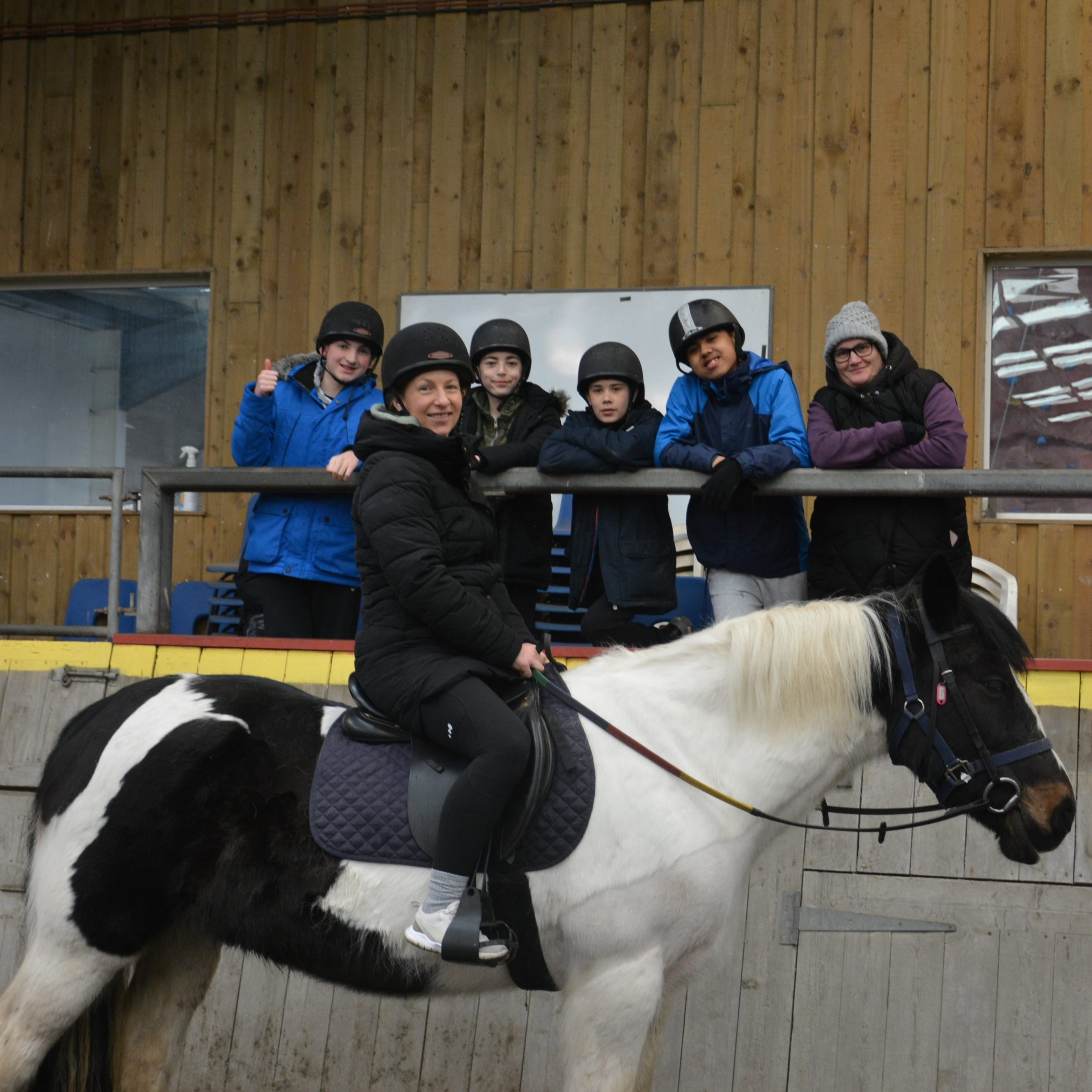 A woman riding black and white horse is sitting side-on but she is smiling at the camera. Behind her, at a higher level, is a group of young people wearing riding hats smiling at the camera.