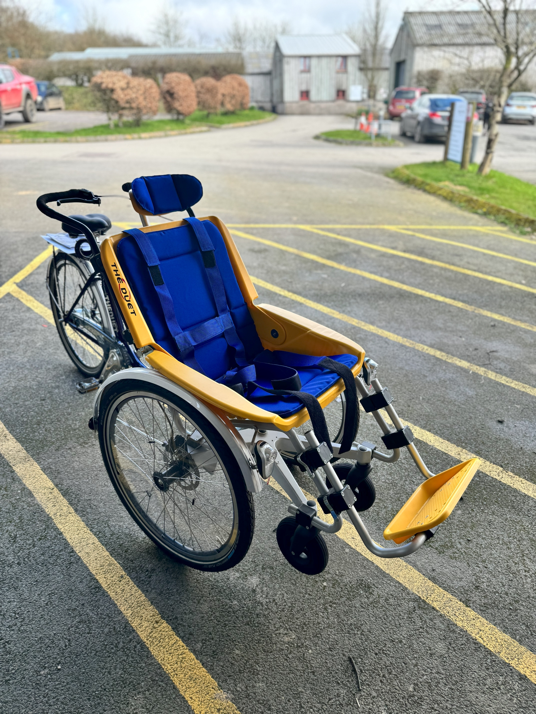 A tricycle with one wheel at the back and two at the front. Atop the two front wheels is a yellow seat with blue cushioning inside, with a footrest at the end of it. There are handlebars behind the seat and a saddle with peddles.