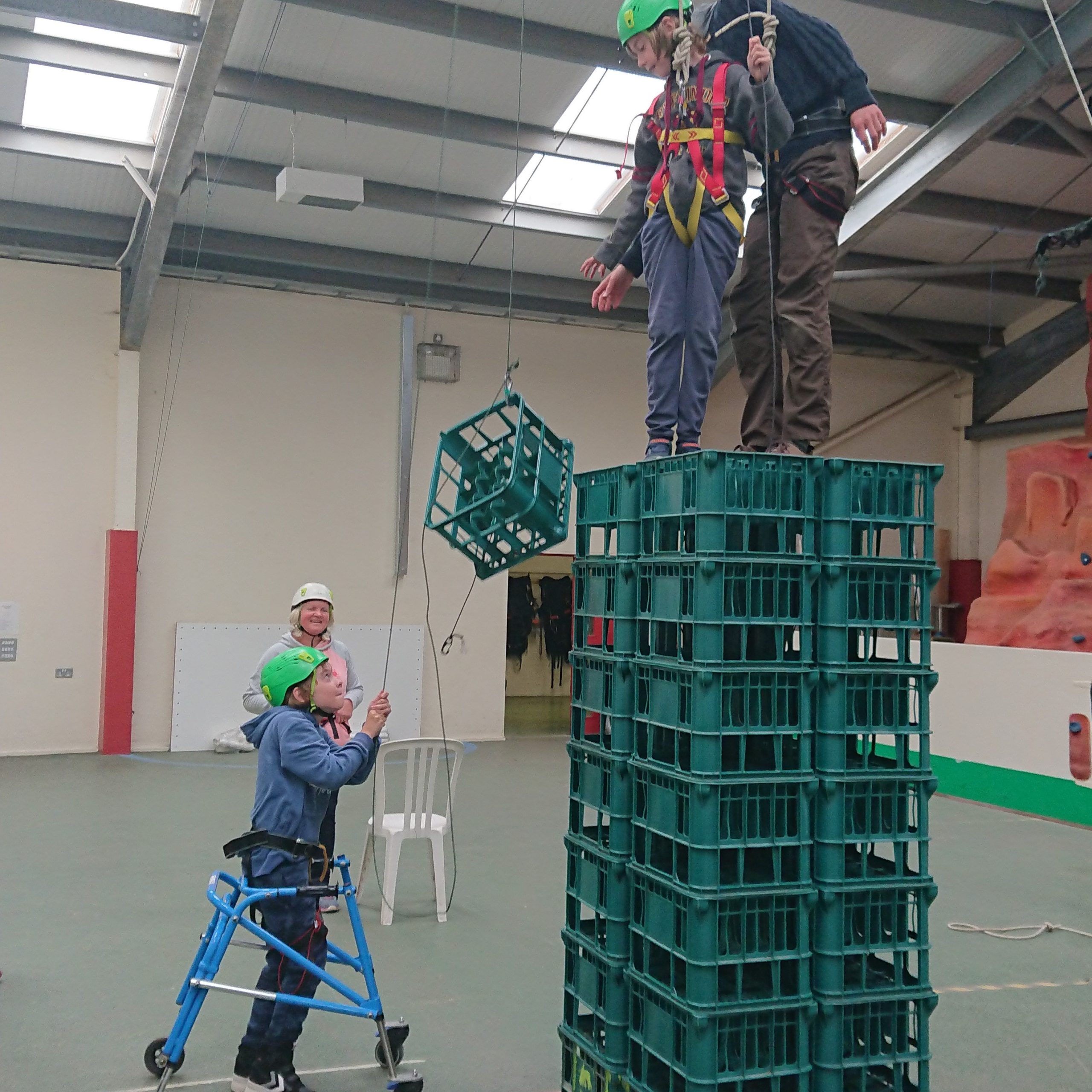 A boy with a blue zimmer frame is standing on the floor of a sports hall, using a small rope to hoist up a crate to two people standing on top of a tower of plastic crates. There is a woman in the background smiling and wearing a white helmet too.