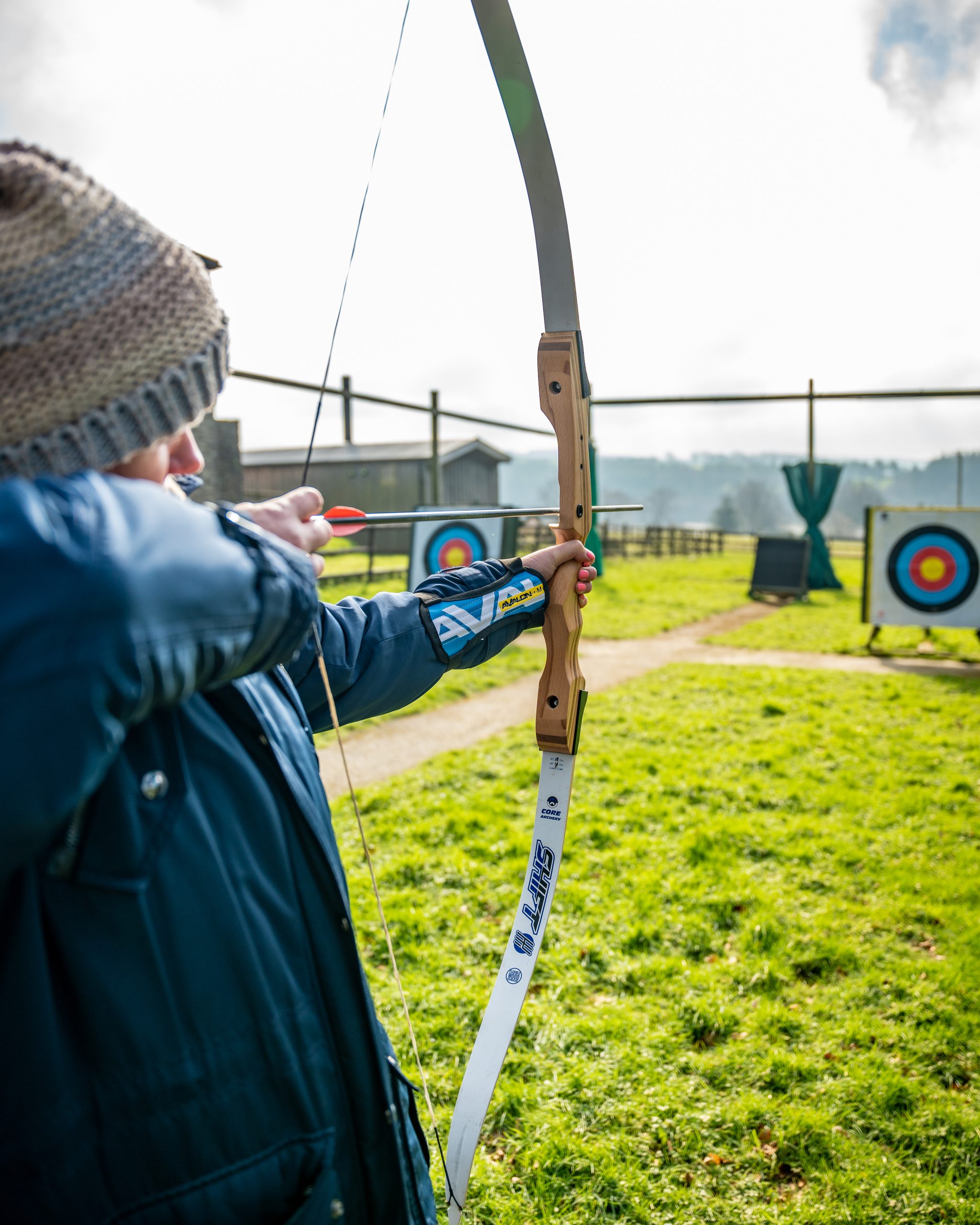 A guest wearing a coat and hat is holding a bow and arrow, with the bowstring drawn back ready to fire and the camera is facing down the line to show where they are aiming: the target.