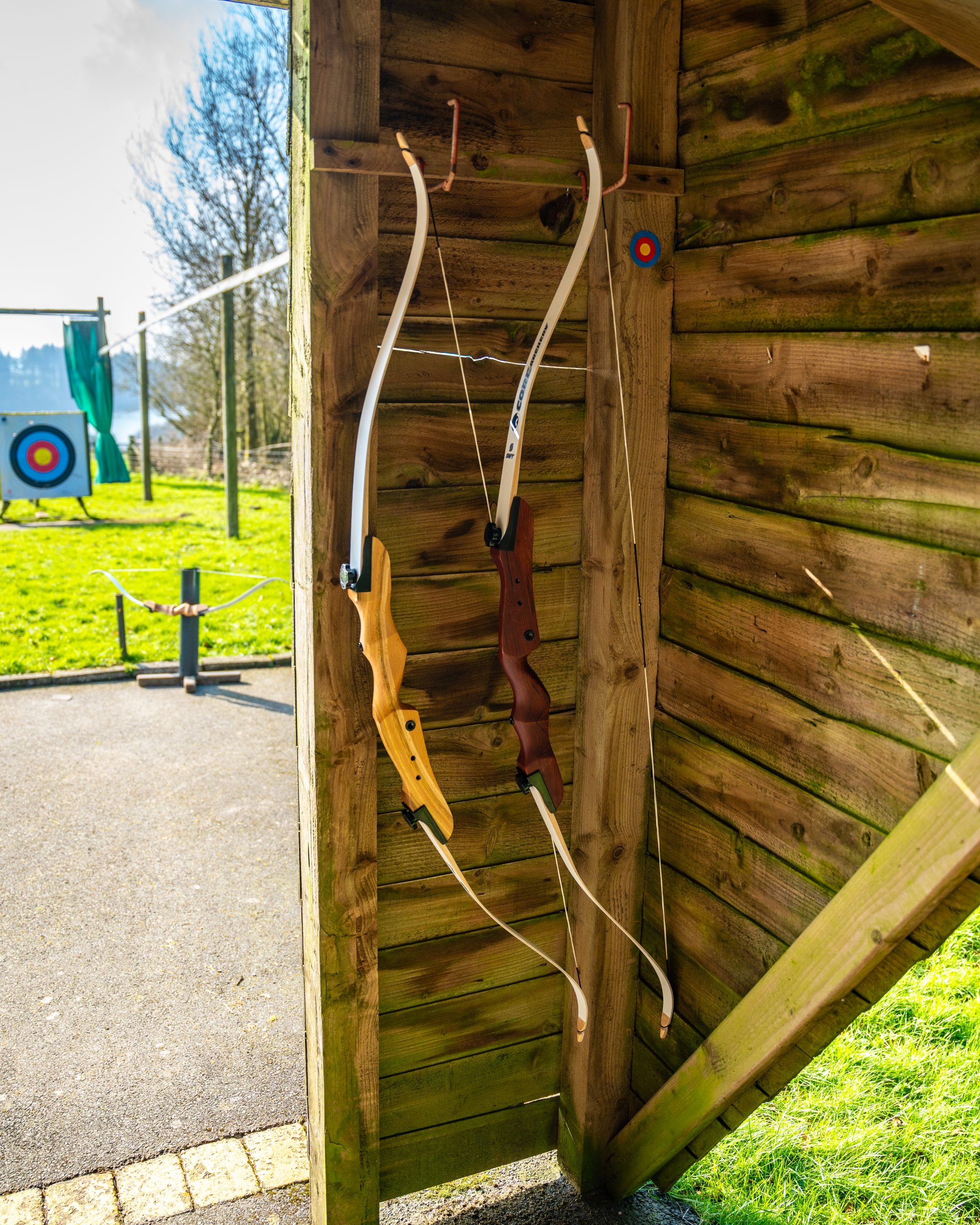 Two large bows with different handle colours are hanging on hooks inside a wood paneled shed. There is a target in the distance, with grass and a paved area behind the shed making up the archery range.