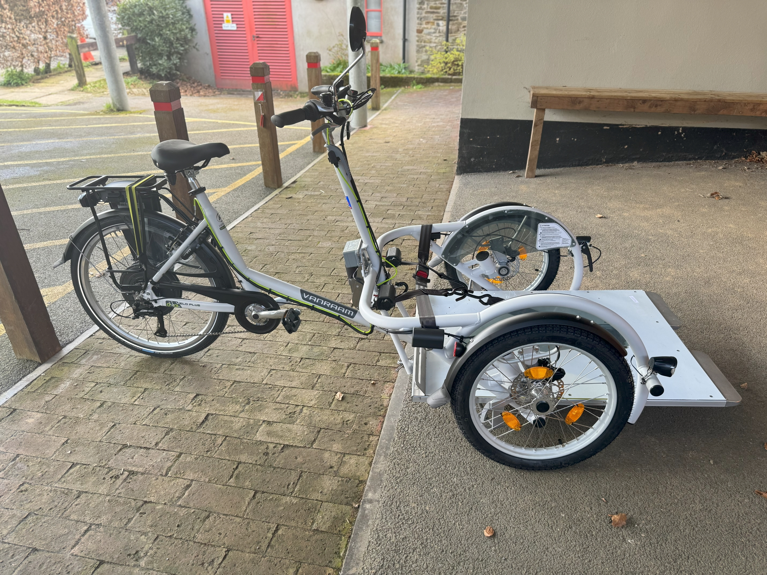 A white tricycle is stationary on a paved area. The front two wheels are either side of a white platform big enough for a wheelchair. The back wheel, the saddle and peddles are like an ordinary bicycle.
