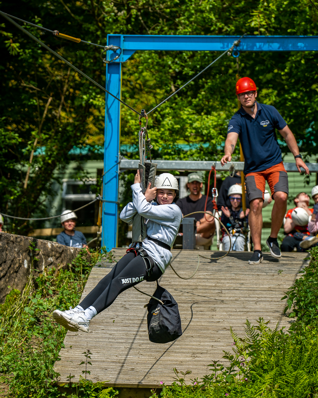 A girl sitting suspended on the Zipwire and moving away from the platform that's behind her, where a member of staff has just released her from the red rope that lets her zoom down the wire. There are other group members in the background watching and clapping.