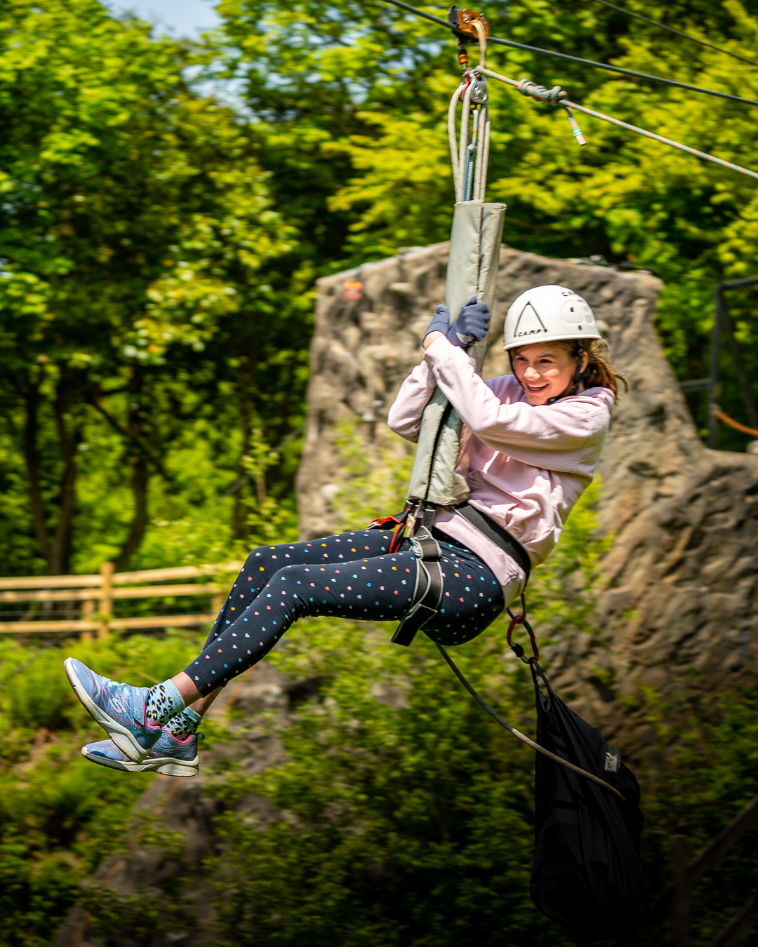 The background is blurry as a girl is zooming along the Zipwire with a big smile on her face, holding onto the cushion that is attached to her and the rope system.