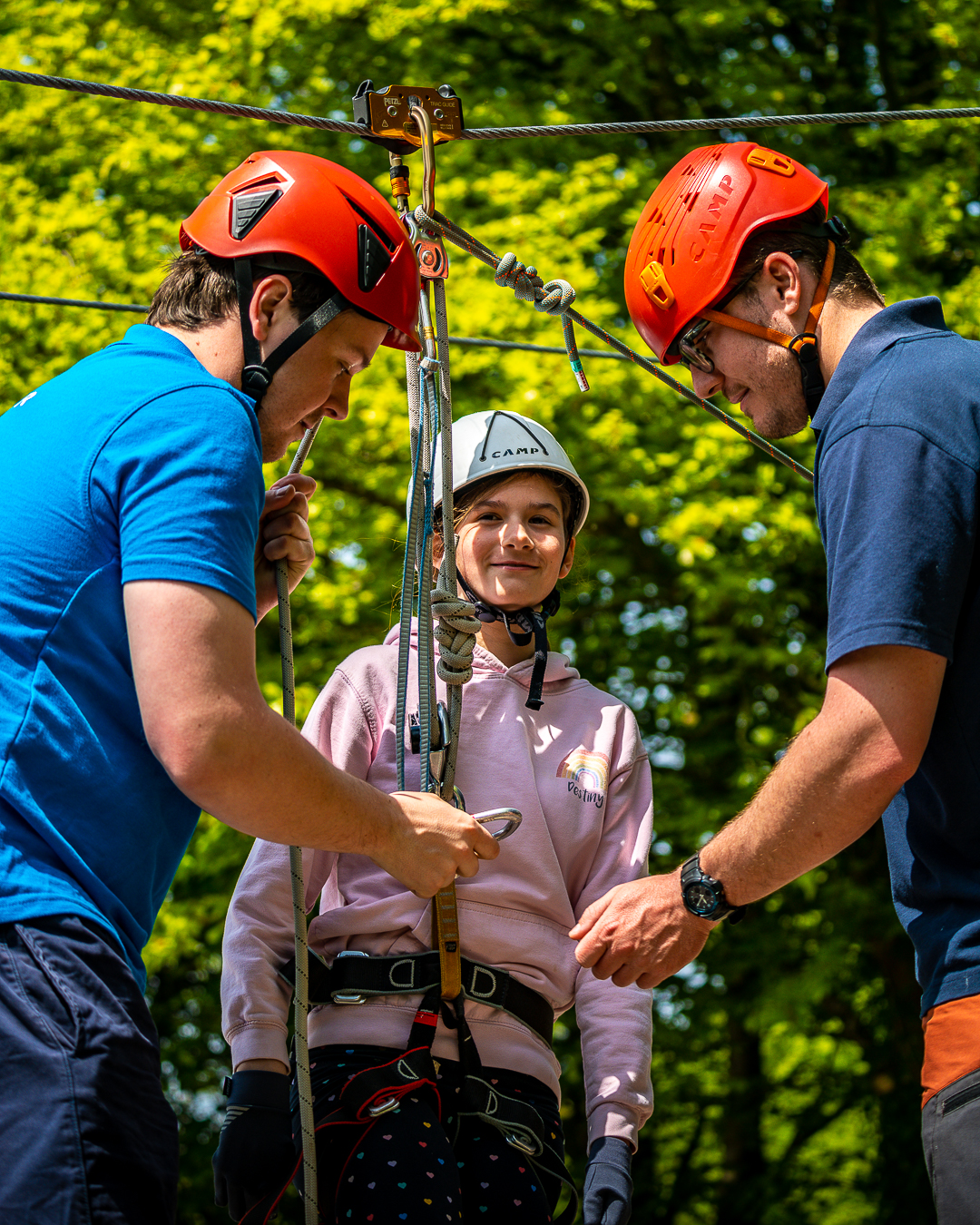 A girl in a pink sweatshirt smiles and looks towards the camera as two men in red helmets are attaching ropes and carabiners to her harness, with a Zipwire above them all.