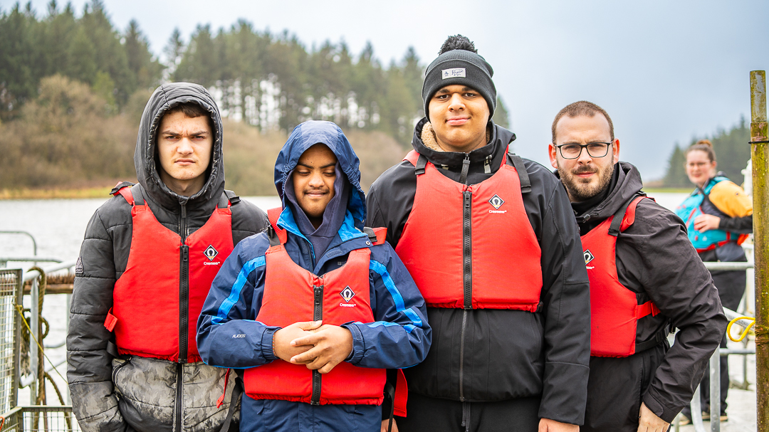 Four guests wearing coats and buoyancy aids are looking at the camera. In the background is a slightly blurred instructor wearing a blue buoyancy aid and in the background of the entire picture is pine forests.
