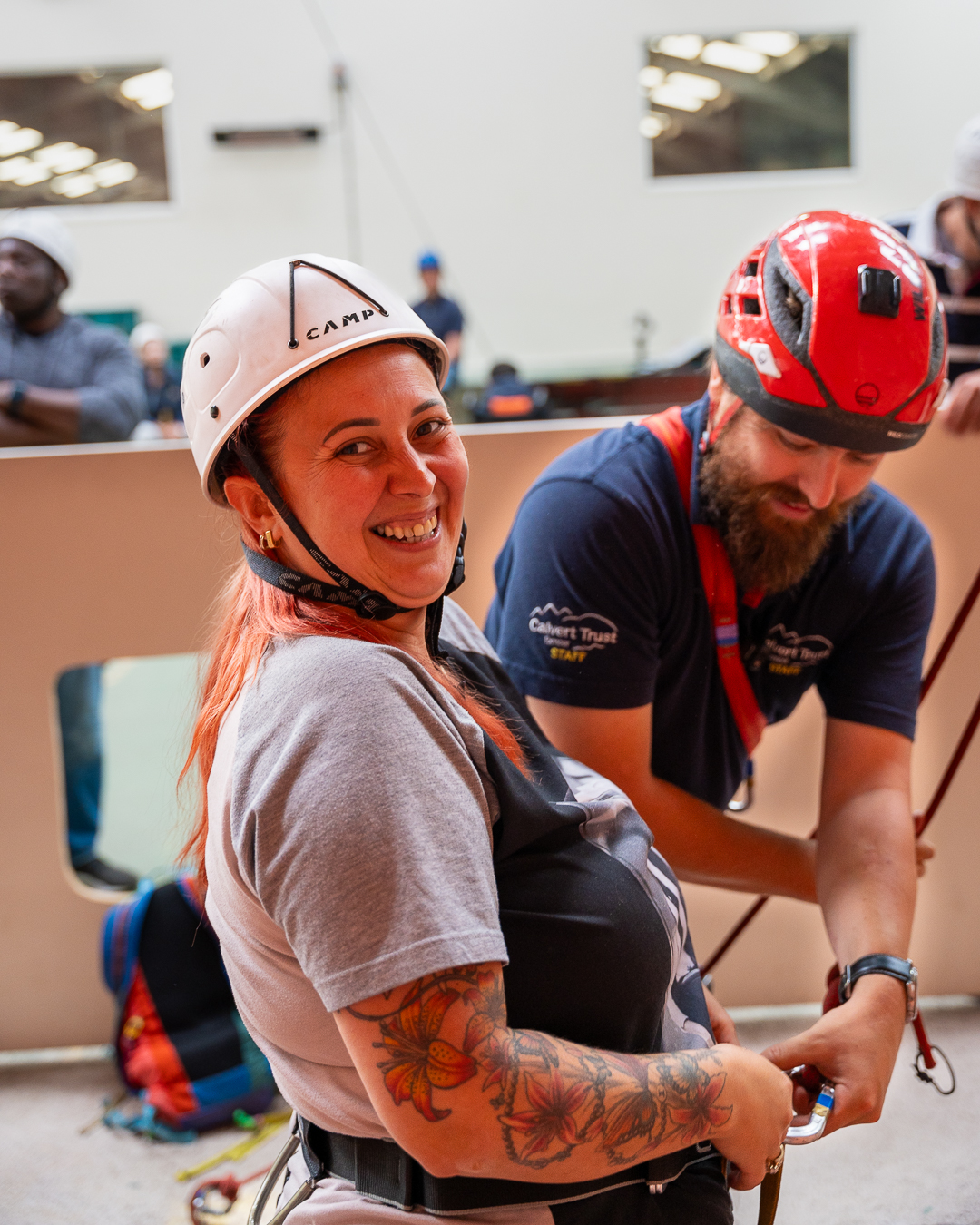 A guest is turning to face the camera, wearing a white helmet and harness, as a member of Calvert Exmoor staff is securing a carabiner to her harness, which is attached to a rope in the indoor climbing wall area.