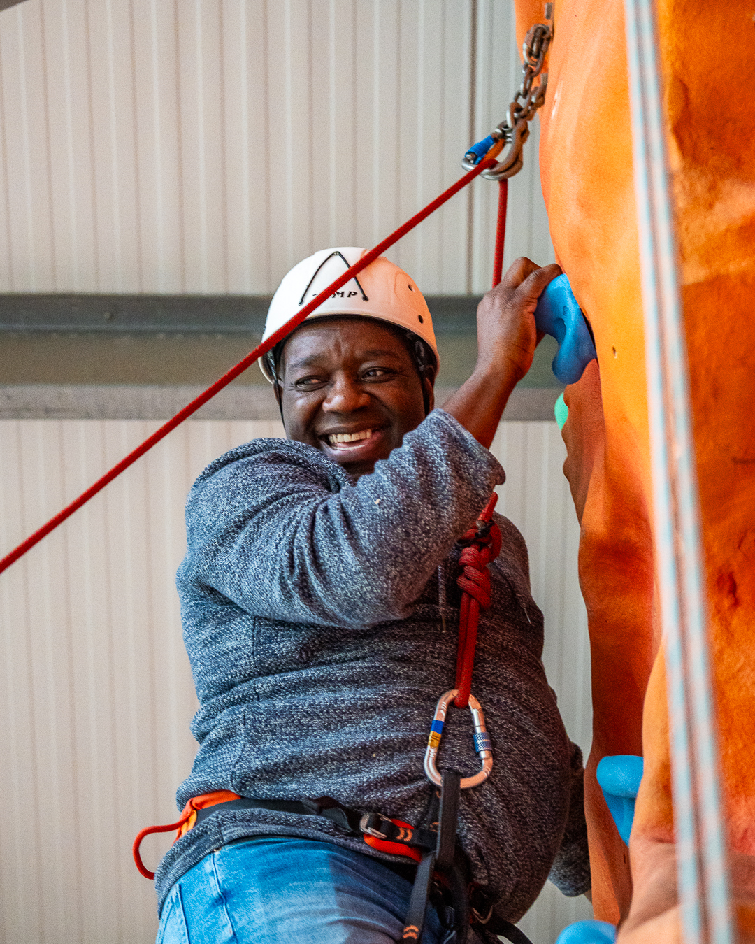 A man is holding a blue hold in an orange indoor climbing wall, with a red rope attached to his harness. He is smiling and looking down at someone out of view, with a white helmet on.