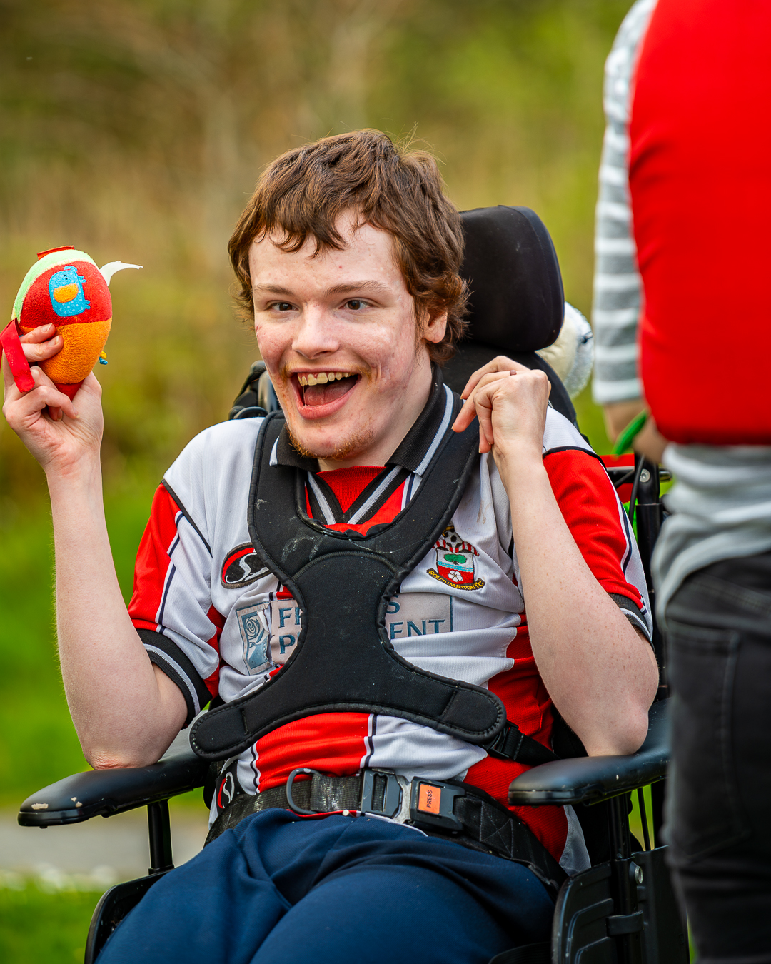 A guest wearing a red and white football shirt is in a wheelchair and smiling with an open mouth towards the camera. He is holding a toy in his hand.