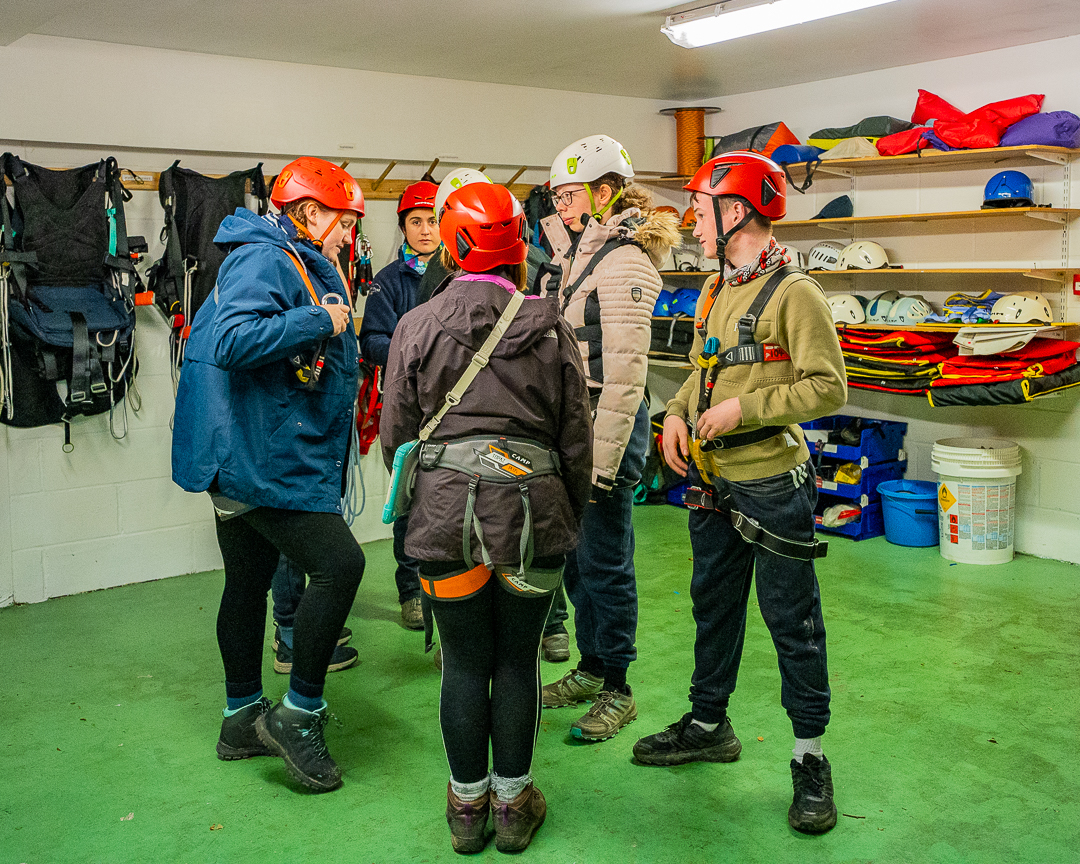 A group of guests are in the equipment store room wearing helmets and harnesses and facing each other. In the background there are shelves with equipment on.