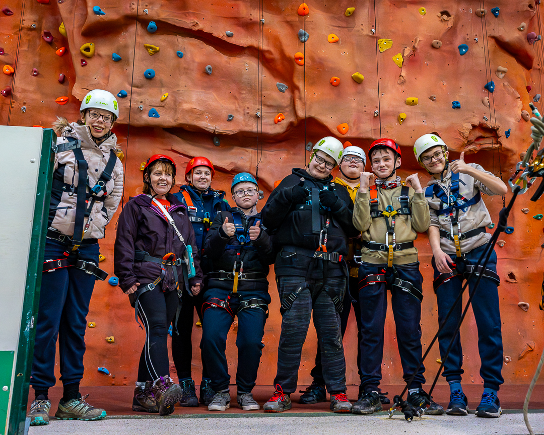 A group of guests wearing helmets and harnesses are standing in front of the inside climbing wall, looking at the camera, smiling and holding their thumbs up.