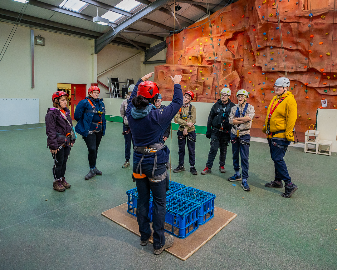 In the background of this photo is the indoor climbing wall, then in the middle there is a group of guests who are wearing harnesses and helmets and facing the camera but looking at a Calvert Exmoor instructor who has their back to the camera, with their arms raised above four blue plastic crates on the floor. The instructor is giving the the guests their introduction to the activity of accessible crate stacking.